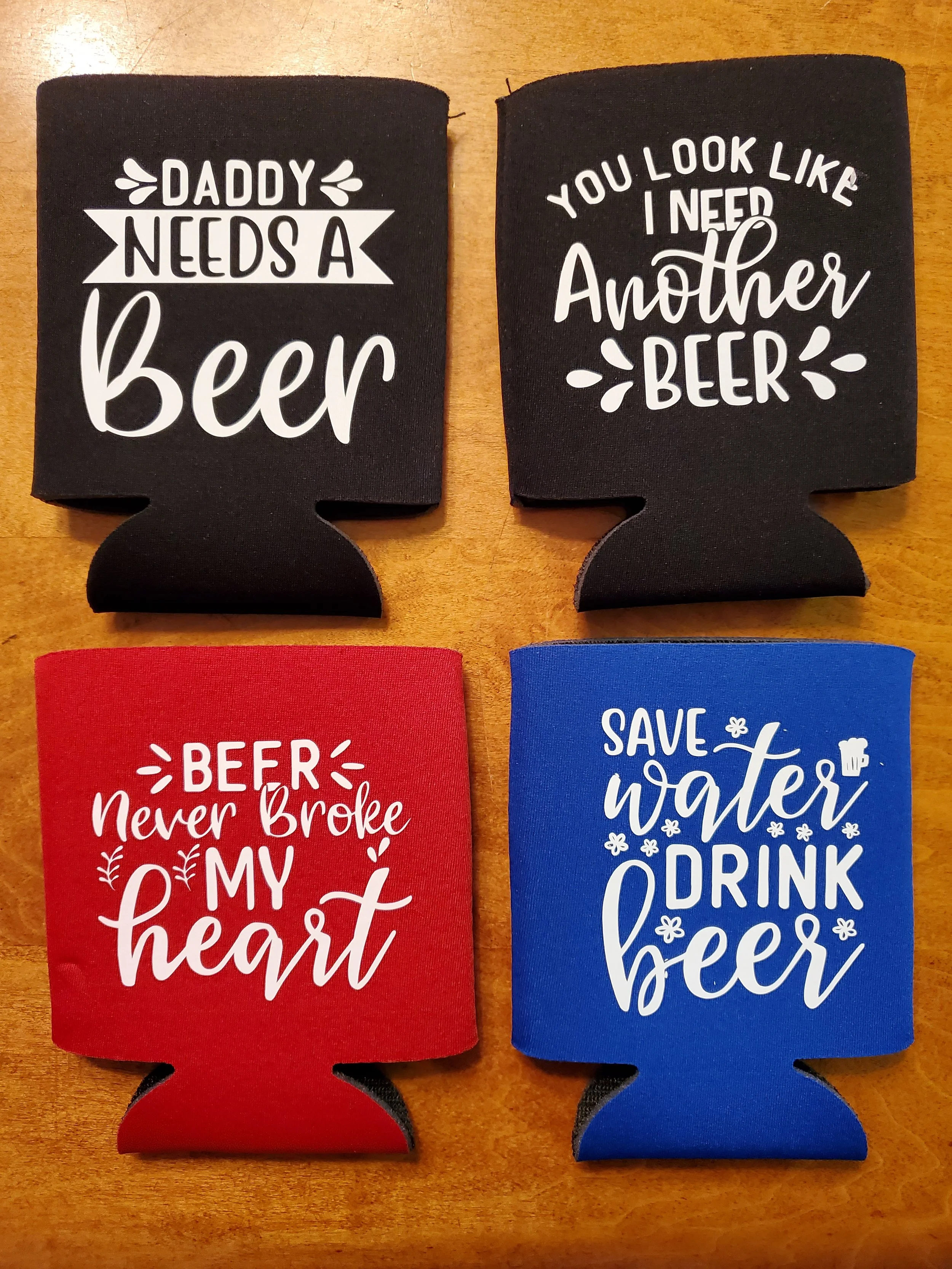 Four koozies with beer-themed sayings on a wooden surface. Top left: "Daddy needs a beer." Top right: "You look like I need another beer." Bottom left: "Beer never broke my heart." Bottom right: "Save water, drink beer," with small beer and flower il