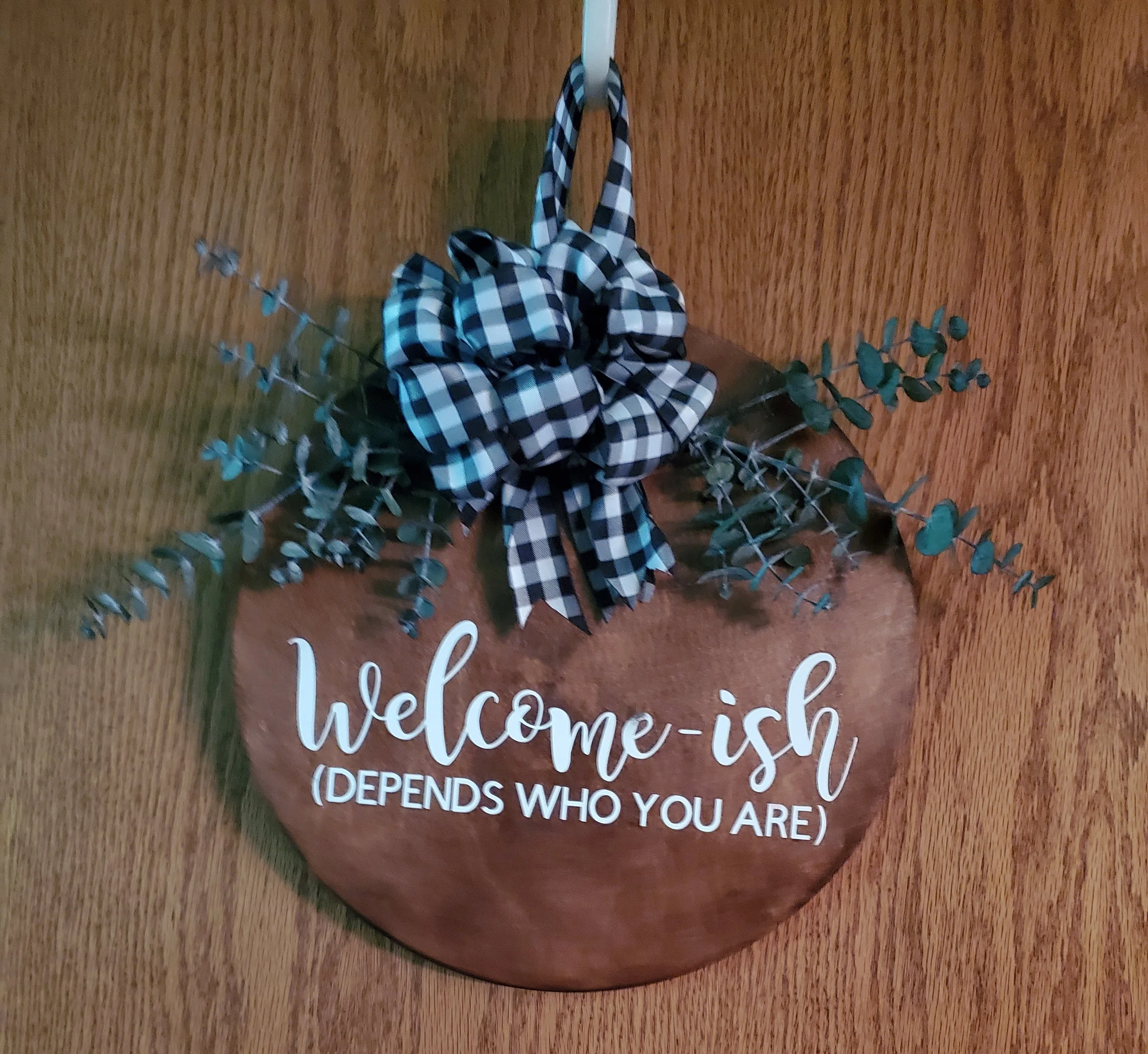 Wooden wall decoration with a black and white checkered bow, faux eucalyptus leaves, and a round wooden sign that reads 'welcome-ish (depends who you are)' in white cursive and block letters.