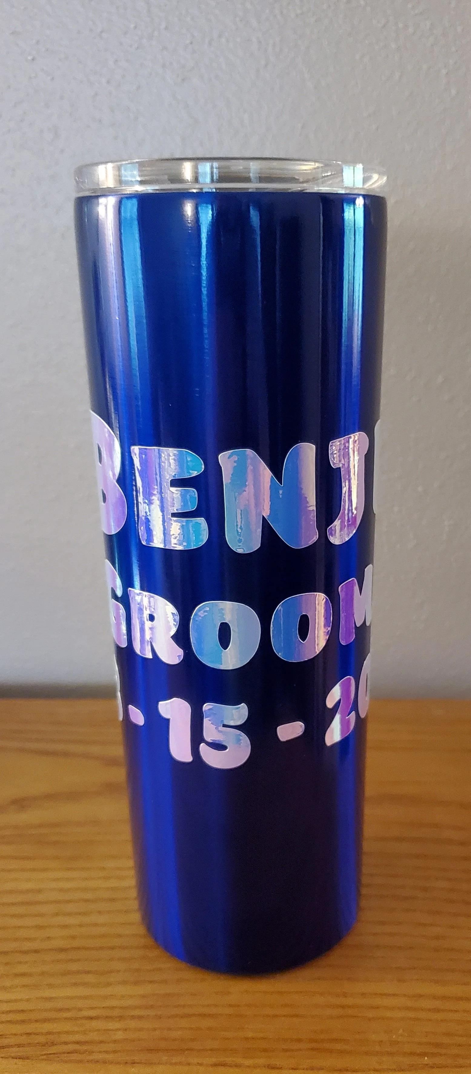 Tall blue tumbler with iridescent writing that says "BENJ" and "15-3" on a wooden surface with a neutral wall background.