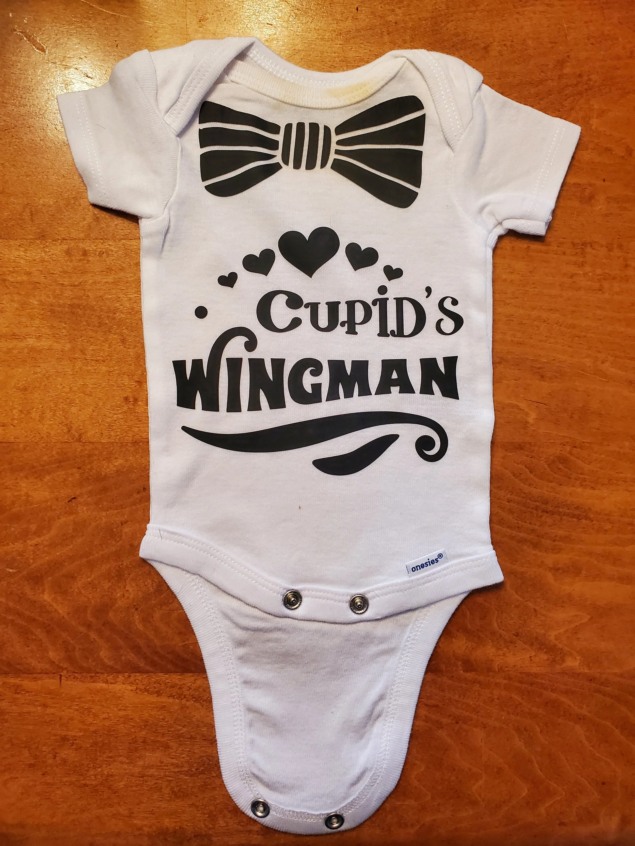 White baby onesie with black graphic print of a bow tie, hearts, and the text 'Cupid's Wingman'.