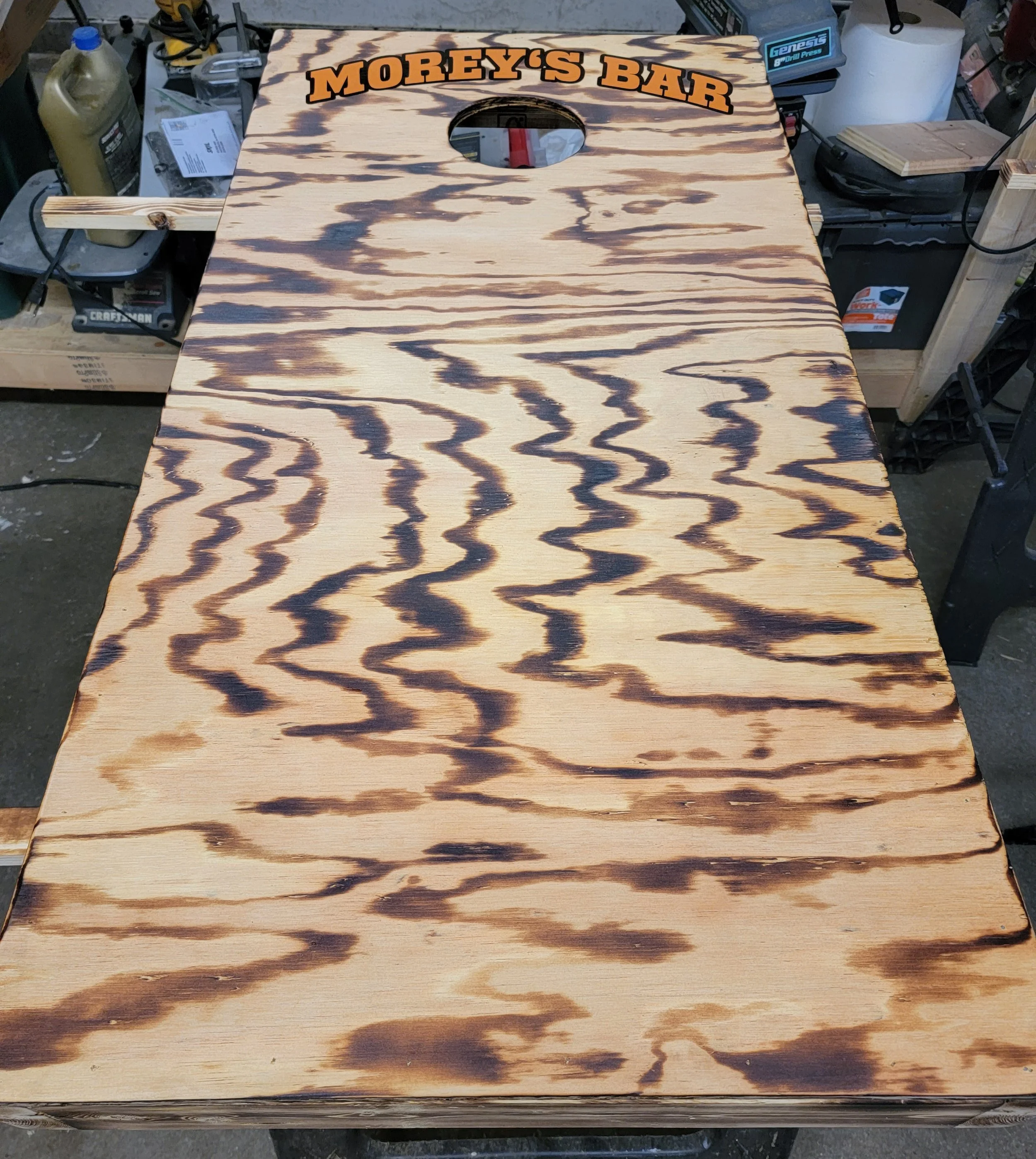 Wooden cornhole game board with burnt design, labeled 'Morey's Bar' at the top, in a workshop setting.