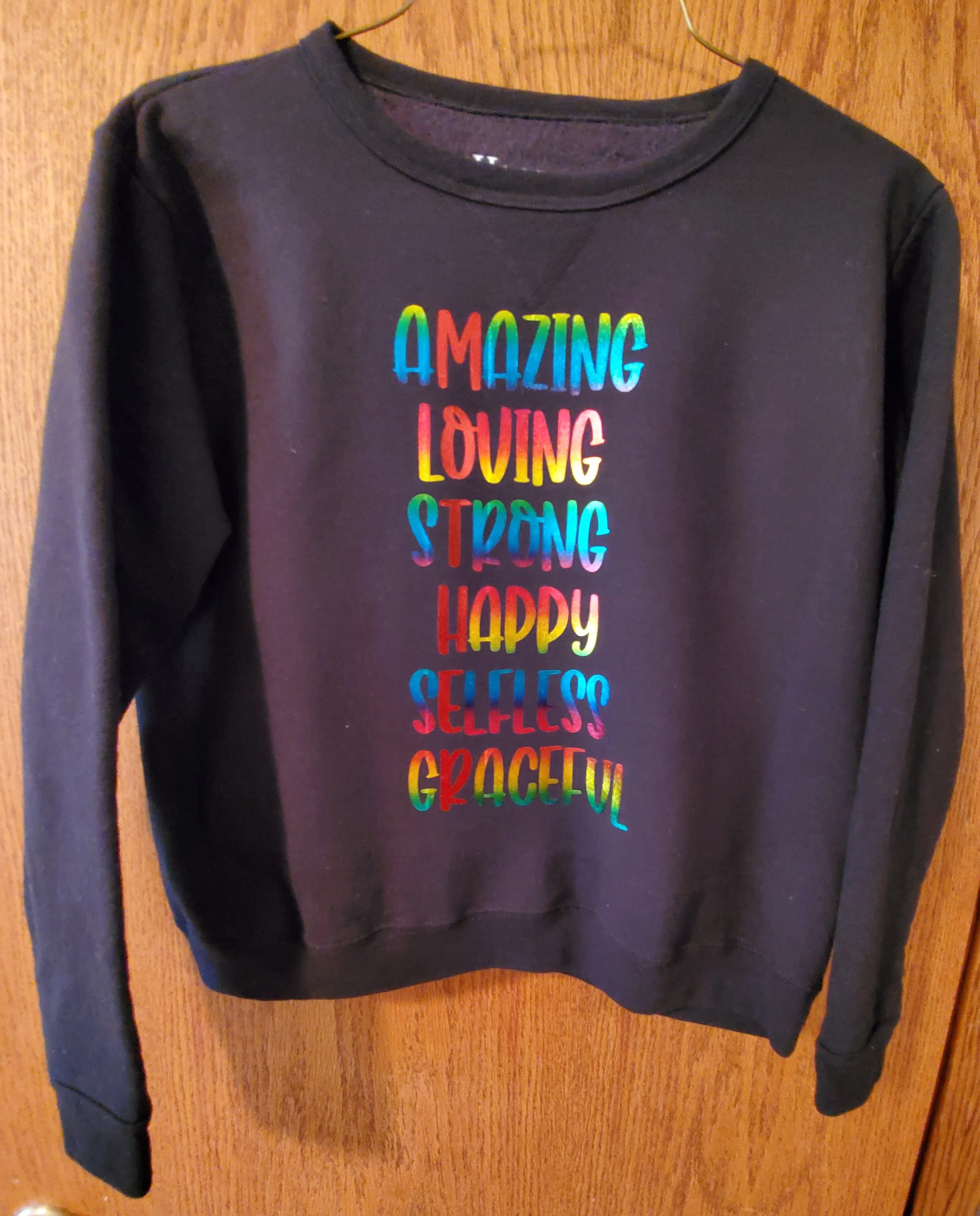 Black sweatshirt hanging on a wooden door with colorful text that reads "AMAZING LOVING STRONG HAPPY SELFLESS GRACEFUL."