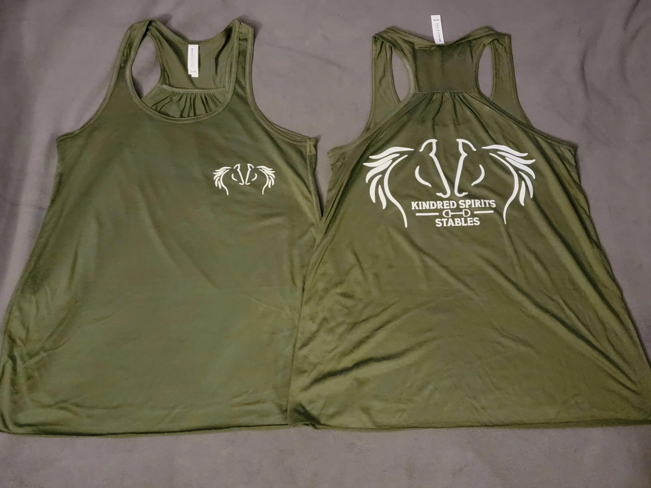 Two olive green tank tops with white graphic designs on the front and back, laid flat on a gray surface.