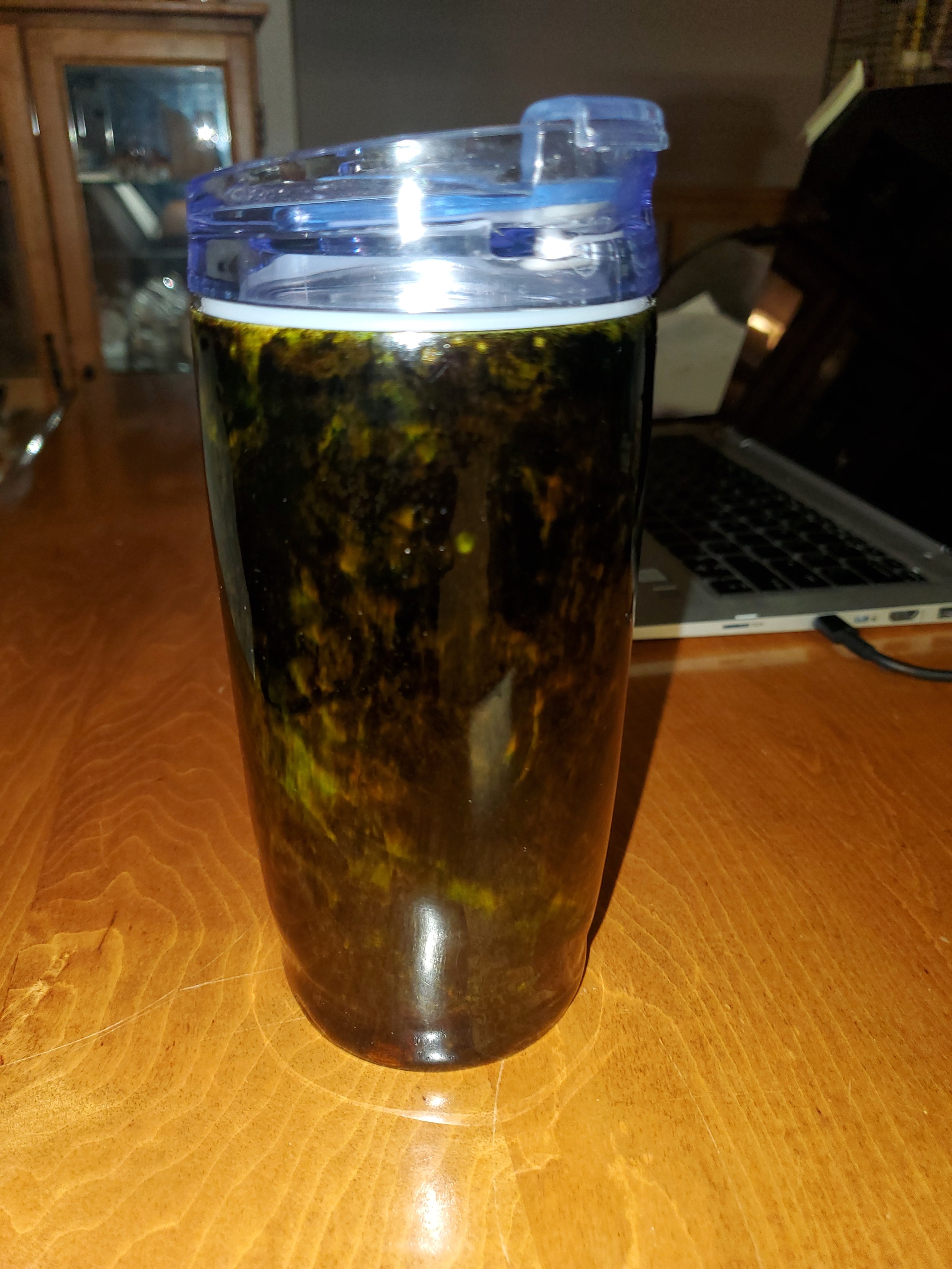 Travel mug with dark liquid, possibly tea or coffee, on wooden table, with a laptop and a china cabinet in the background.