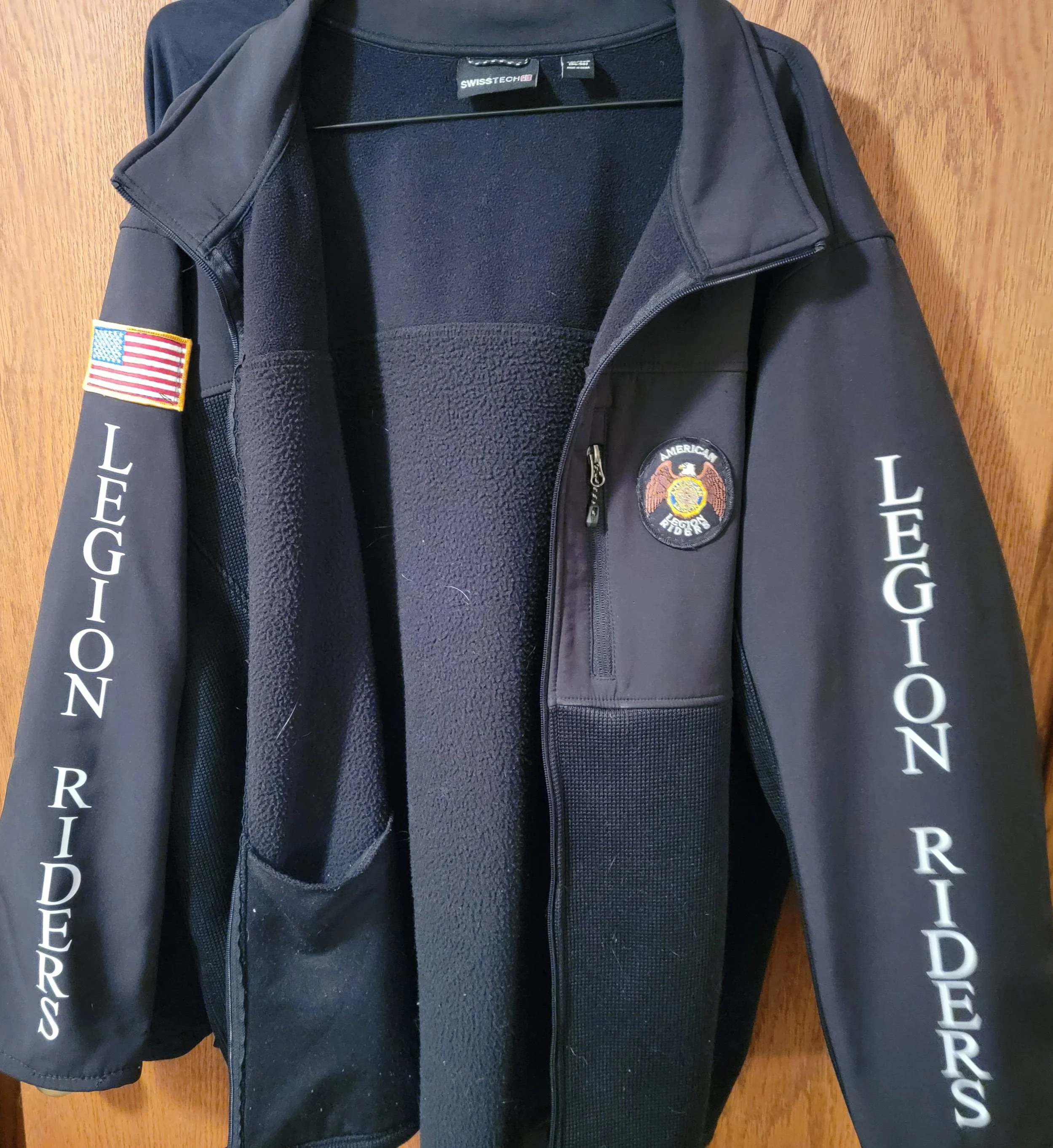 Black jacket with "LEGION RIDERS" printed on the sleeves, a United States flag patch on the shoulder, and a circular emblem patch on the chest, hanging on a wooden background.