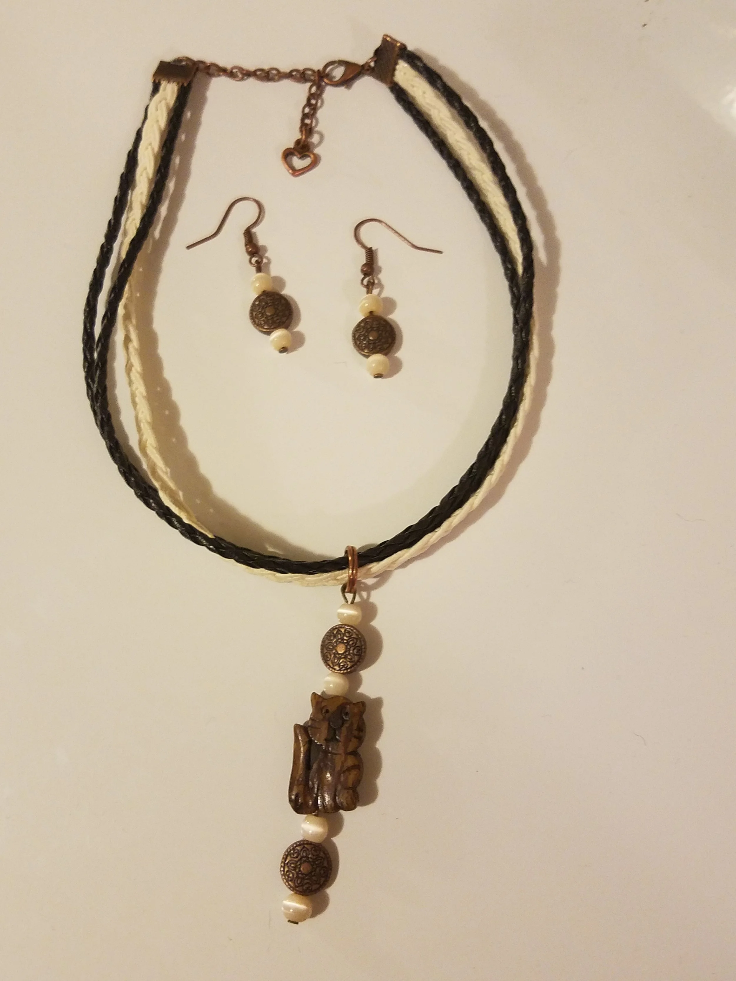 A jewelry set with a necklace and matching earrings hanging on a wall. The necklace features a carved animal pendant, possibly a bear, with beads, and the earrings have similar round beads. The jewelry is coordinated with black and cream cords.