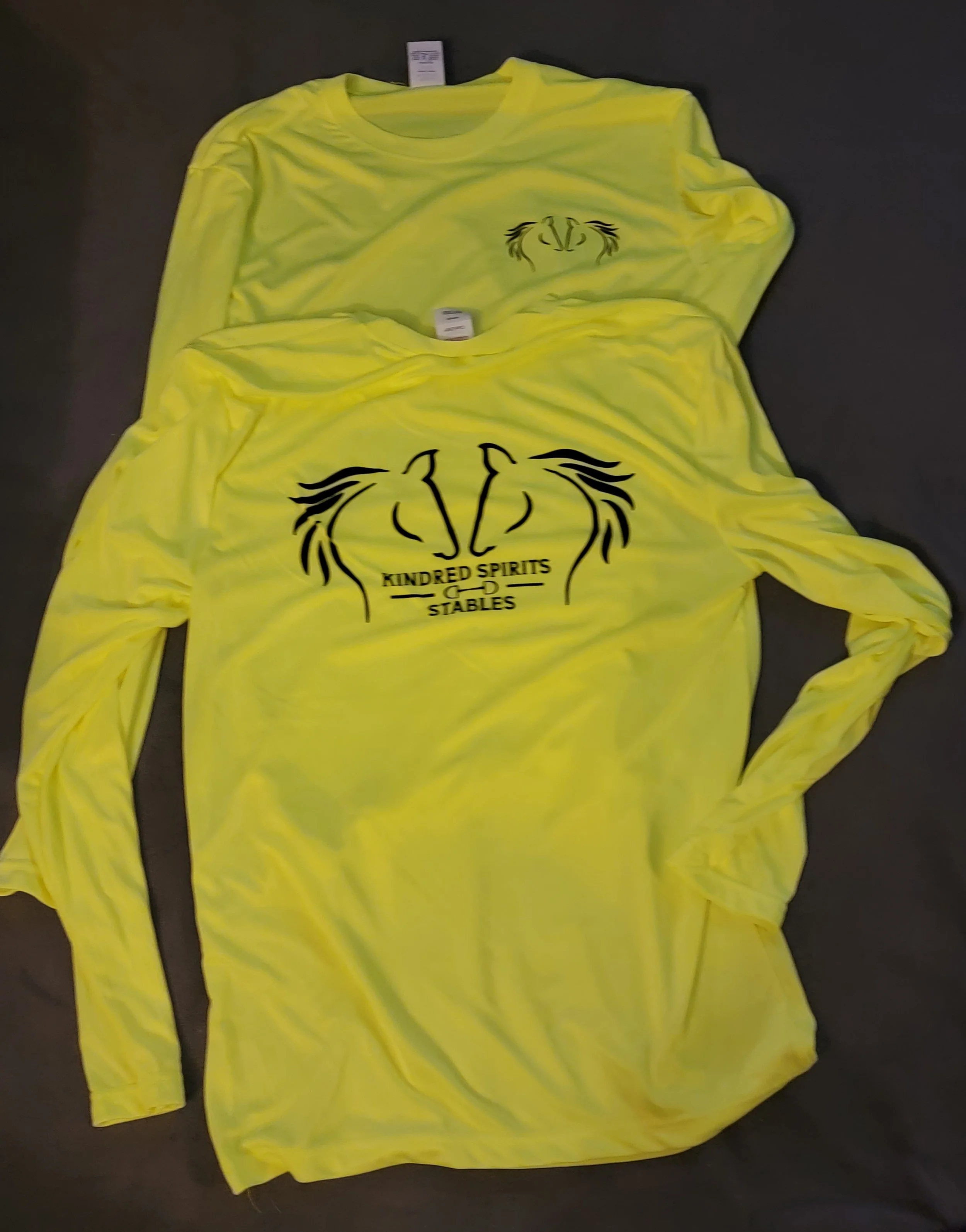Two bright yellow long-sleeve shirts with a black outline of two horses' heads and the text "KINDRED SPIRITS STABLES" on the front. They are laid on a flat dark surface.