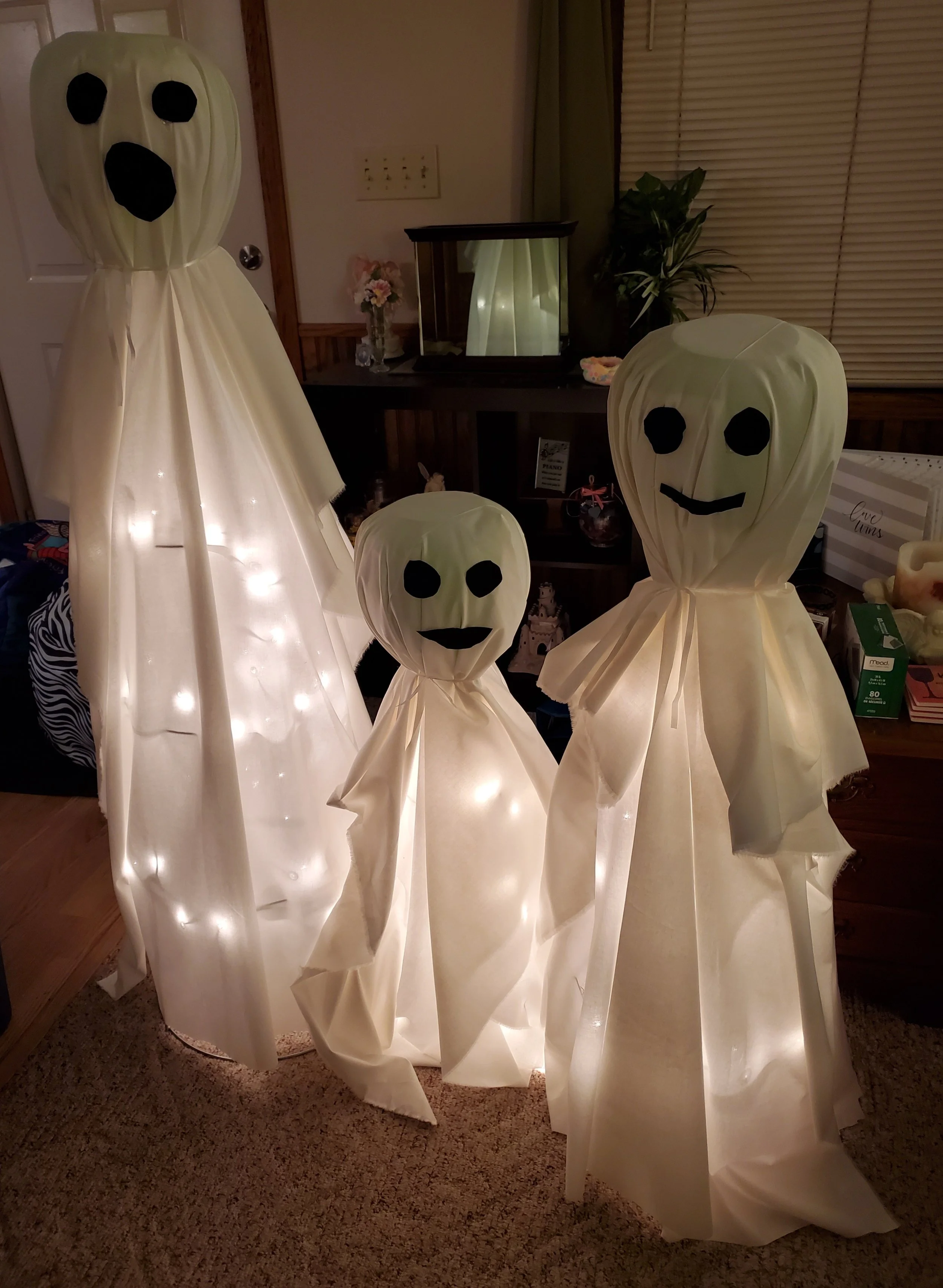 Three ghost-shaped Halloween lanterns with smiling faces and black eyes, illuminated from within and placed indoors. One larger ghost is on the left, with a tall, flowing white sheet. The other two are smaller, one on the right with a smiling face an