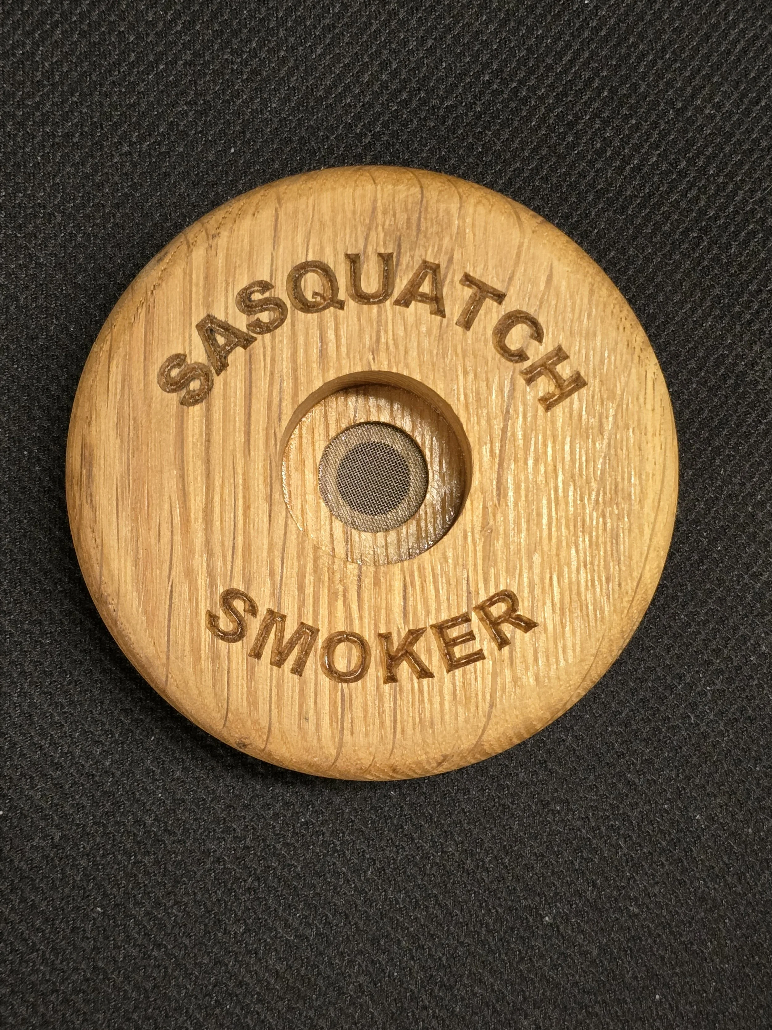 Round wooden item with engraved text 'SQUATCH' on the top and 'SMOKER' on the bottom, with a small mesh vent in the center. 