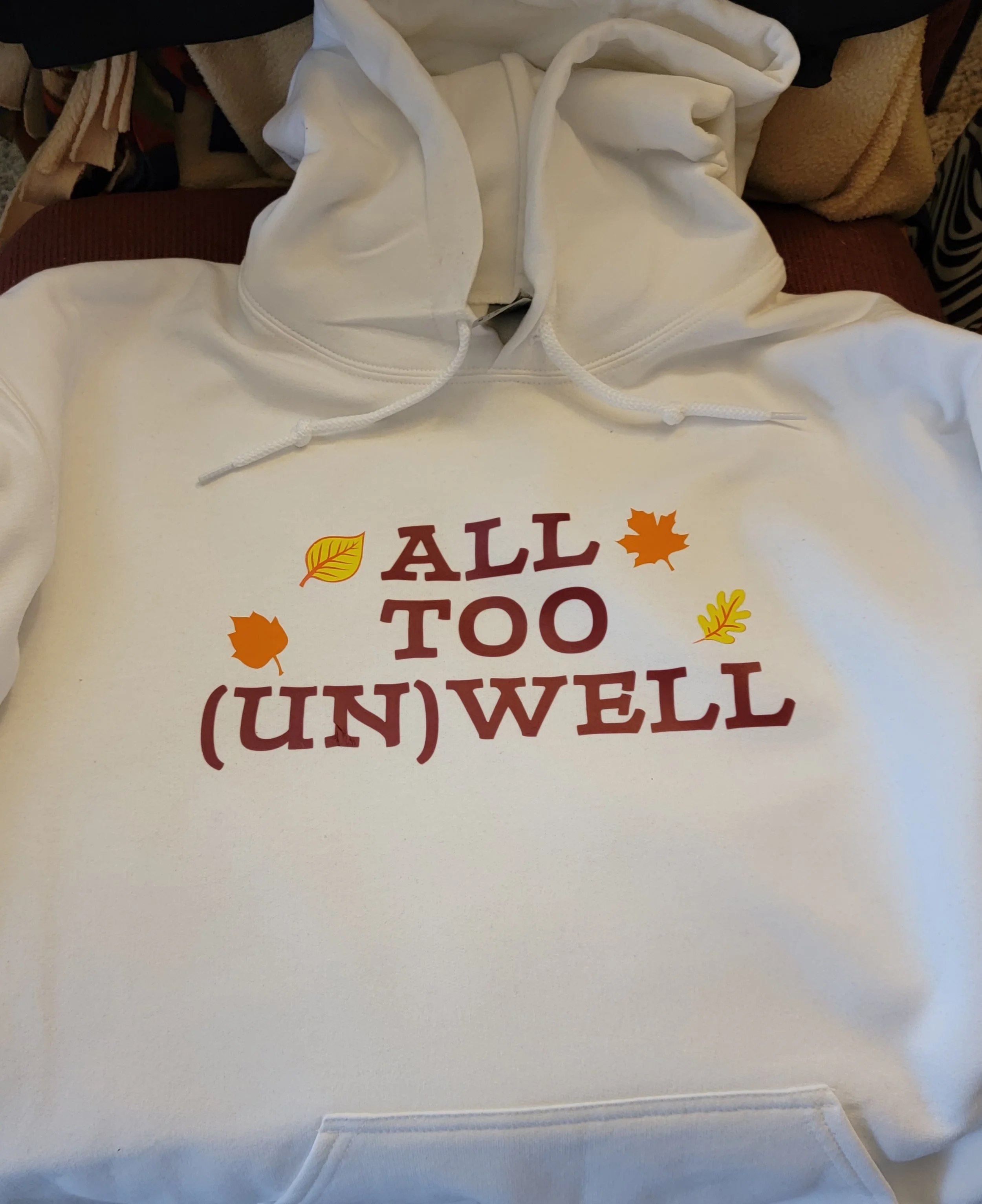 White hoodie with the phrase 'ALL TOO (UN)WELL' in red and brown letters, decorated with yellow and orange fall leaves.