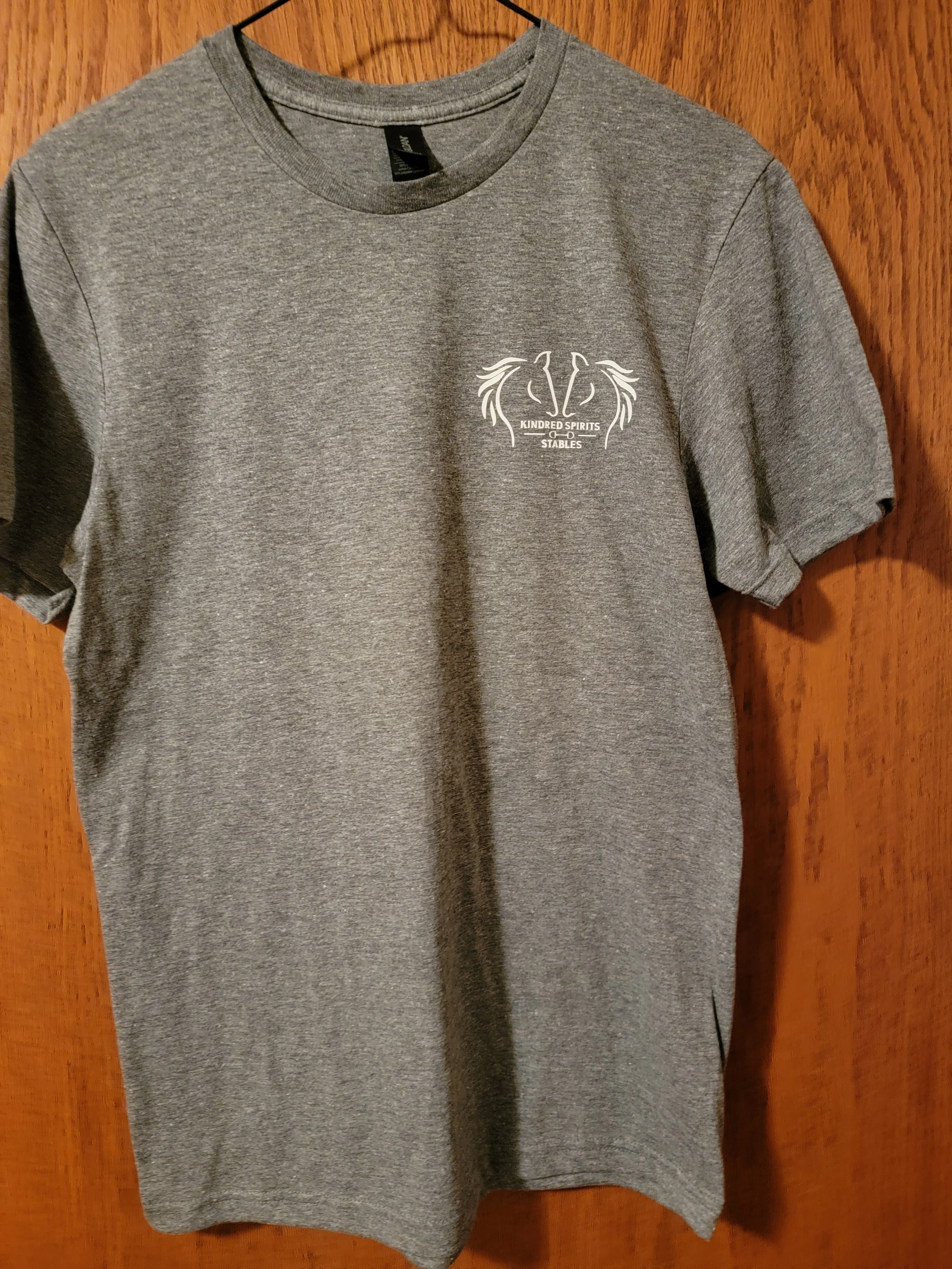 A gray t-shirt hanging on a hanger against a wooden background, with a small white logo on the chest reading 'KINDRED SPIRITS STABLES'.