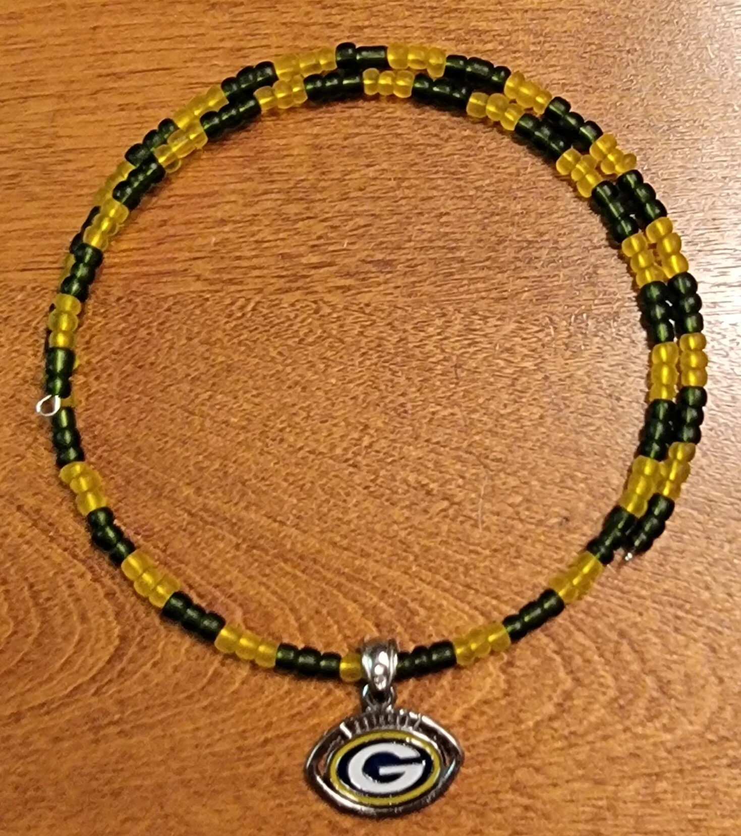 Green and yellow beaded necklace with a small metallic pendant featuring the Green Bay Packers logo, placed on a wooden surface.