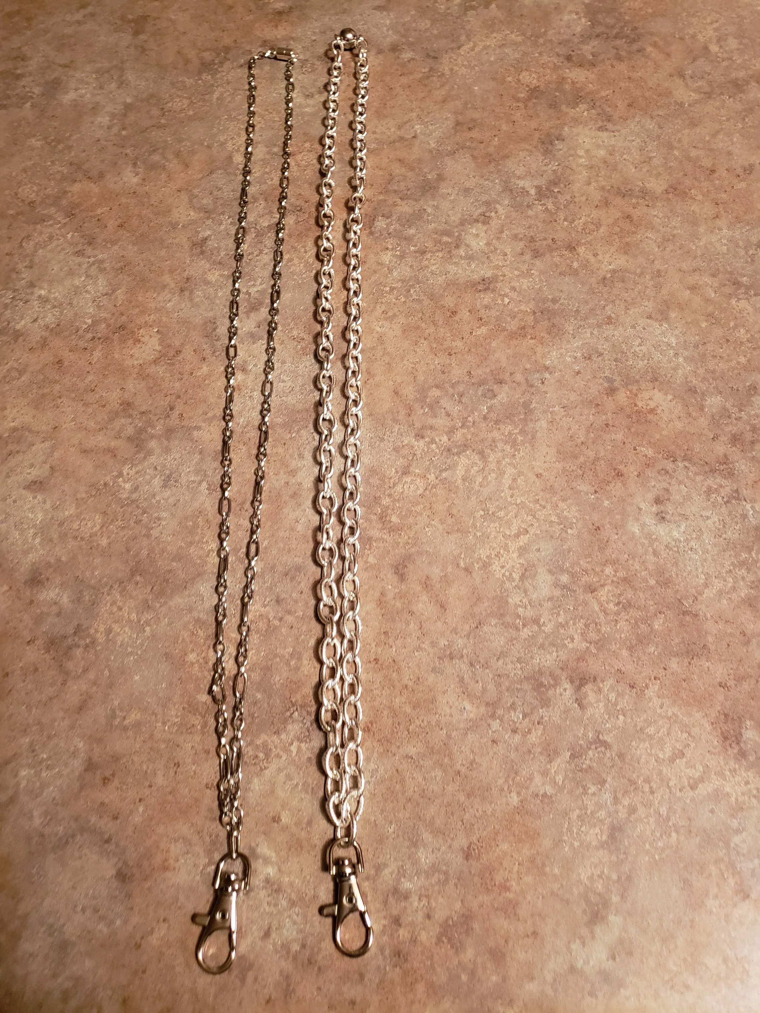 Two silver chains with clasps, one thin and one thick, laid out on a textured brown surface.