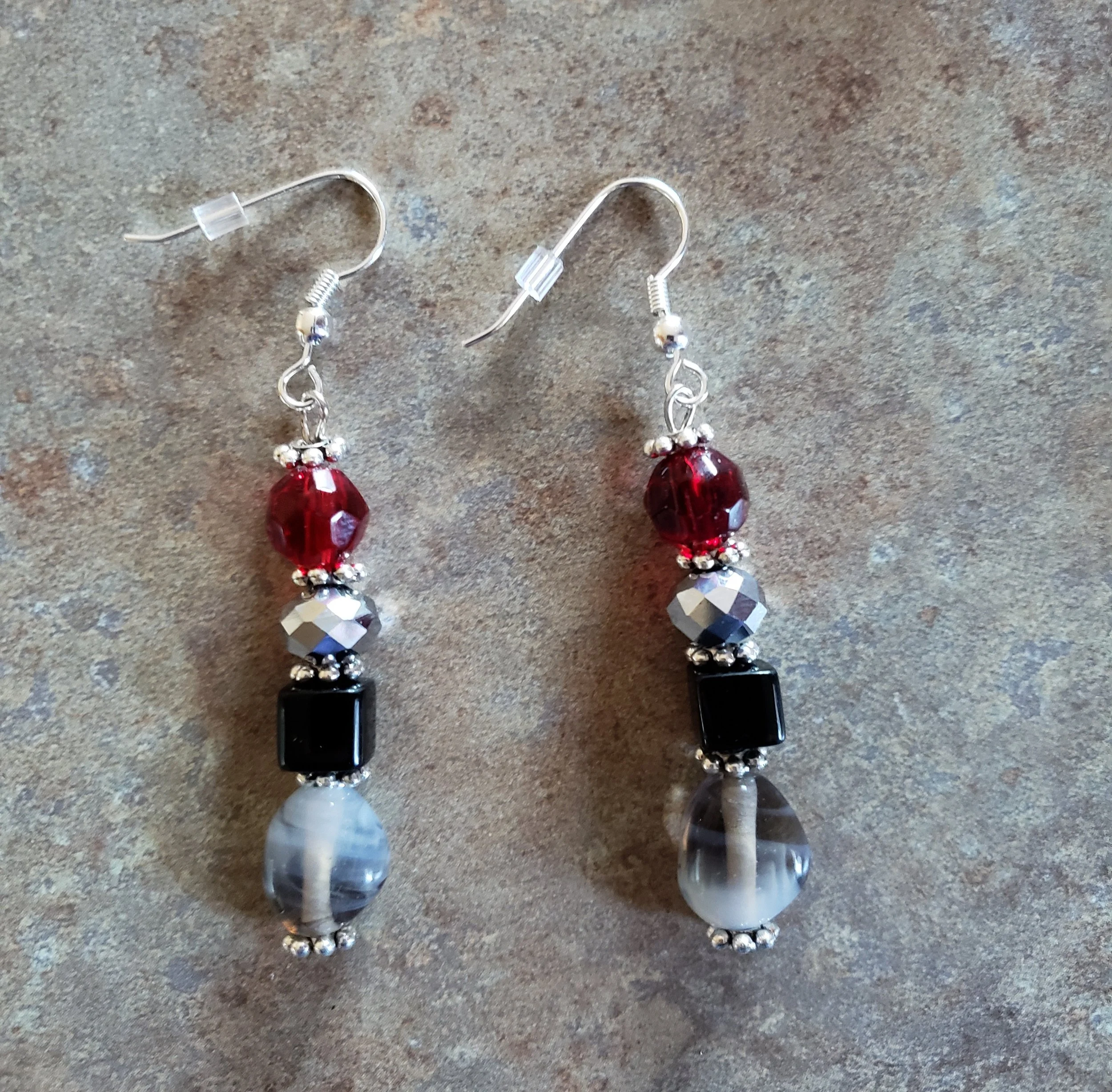 Pair of dangling earrings with various beads including red, black, silver, and gray on a textured surface.