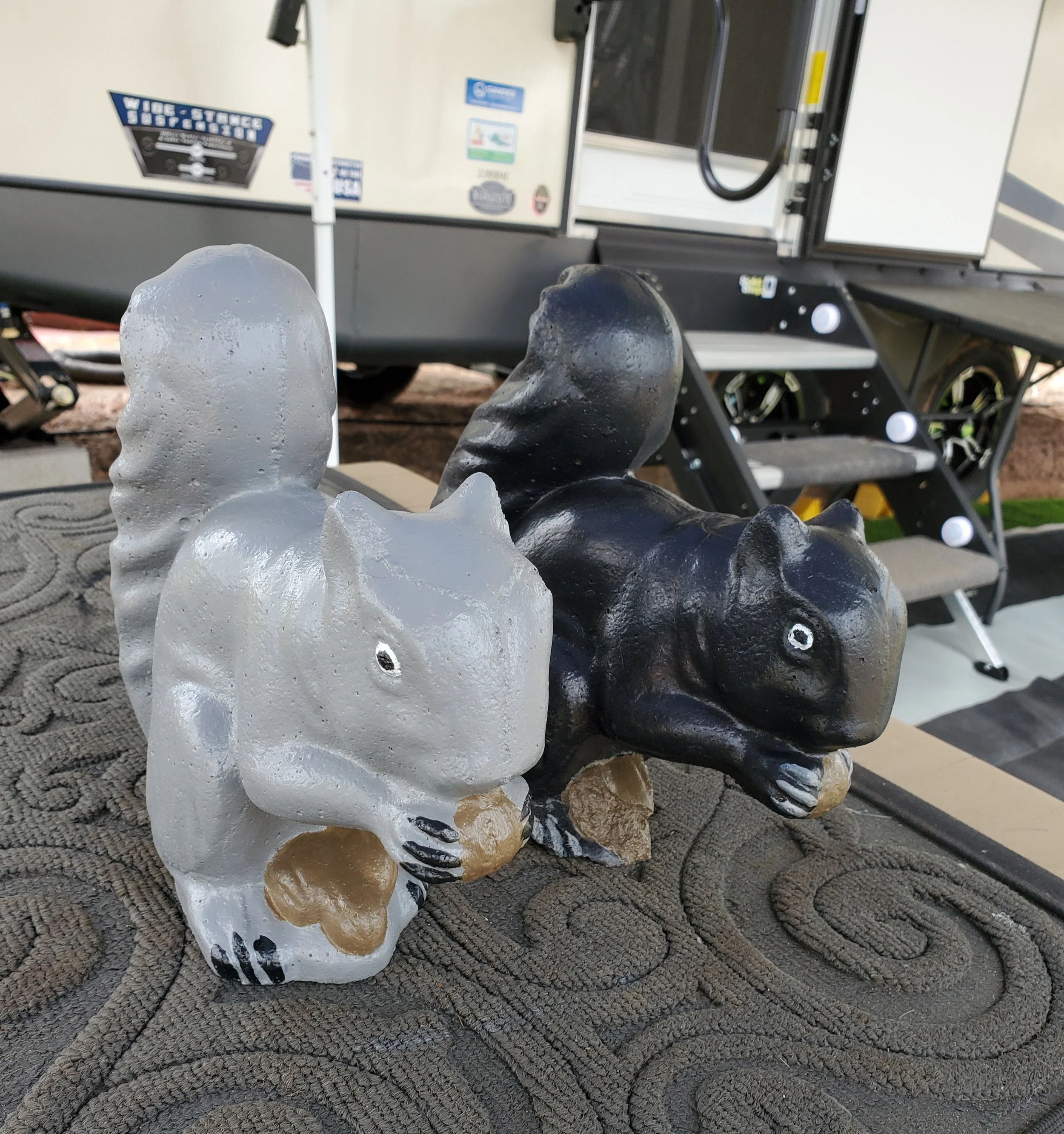 Two squirrel-shaped statues, one painted white and the other black, holding nuts, placed on a textured doormat in front of a trailer or RV with steps.