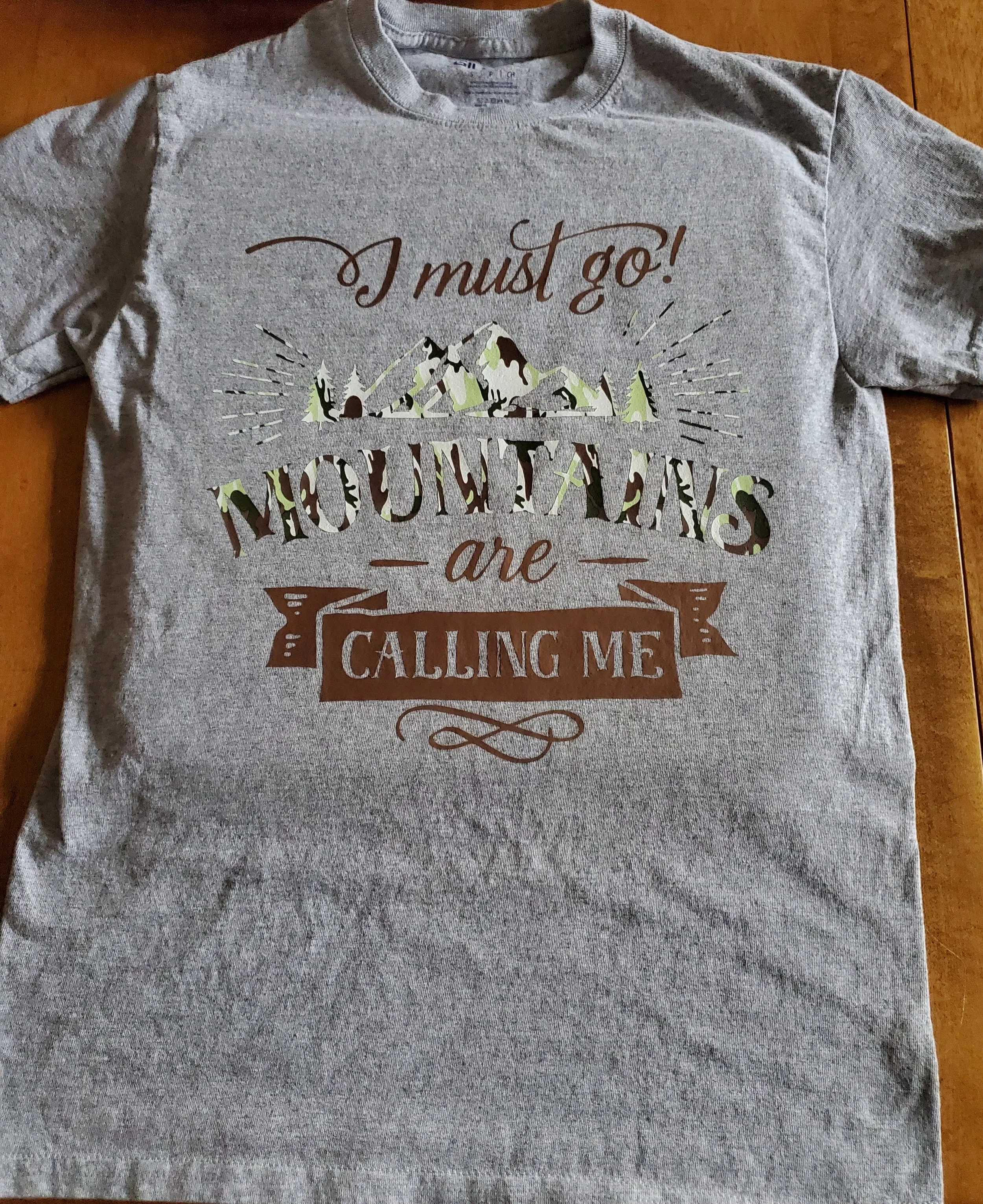 Gray T-shirt with a mountain and pine trees graphic and the text "I must go! MOUNTAINS are CALLING ME" in brown and green lettering.