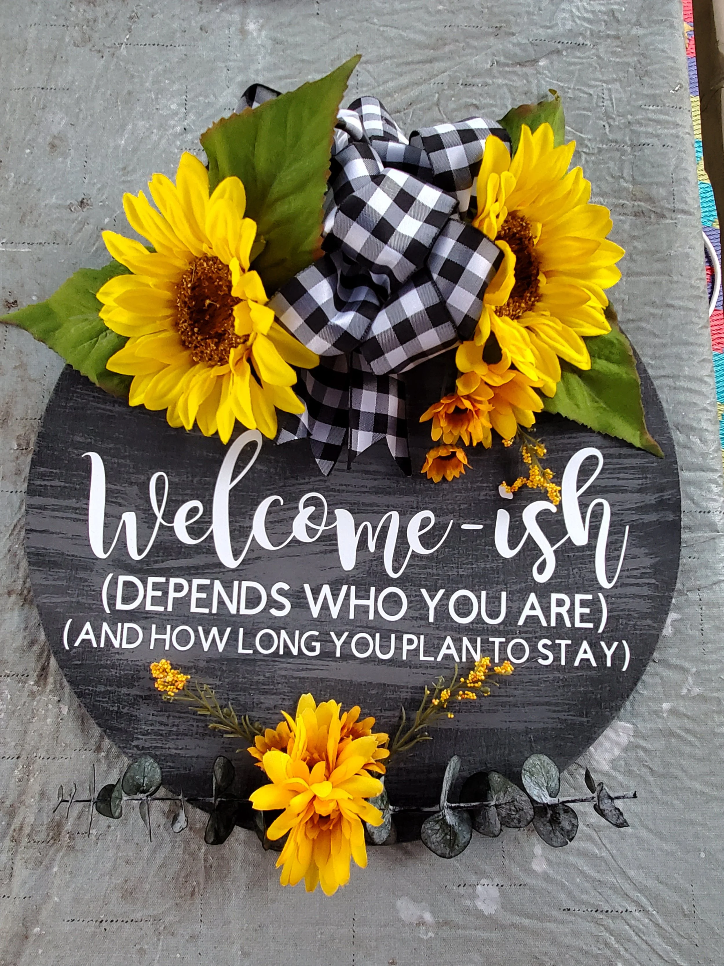 Decorative round sign with sunflowers, leaves, a black and white checkered ribbon, and yellow flowers, displaying the message: 'welcome-ish (depends who you are) (and how long you plan to stay).'