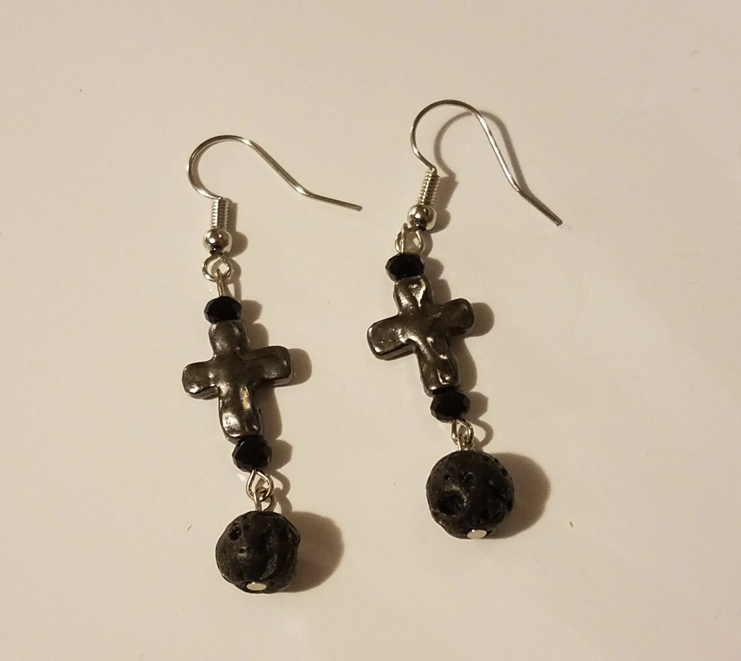 A pair of dangling earrings with silver cross charms, black beads, and round black volcanic beads, on a white background.