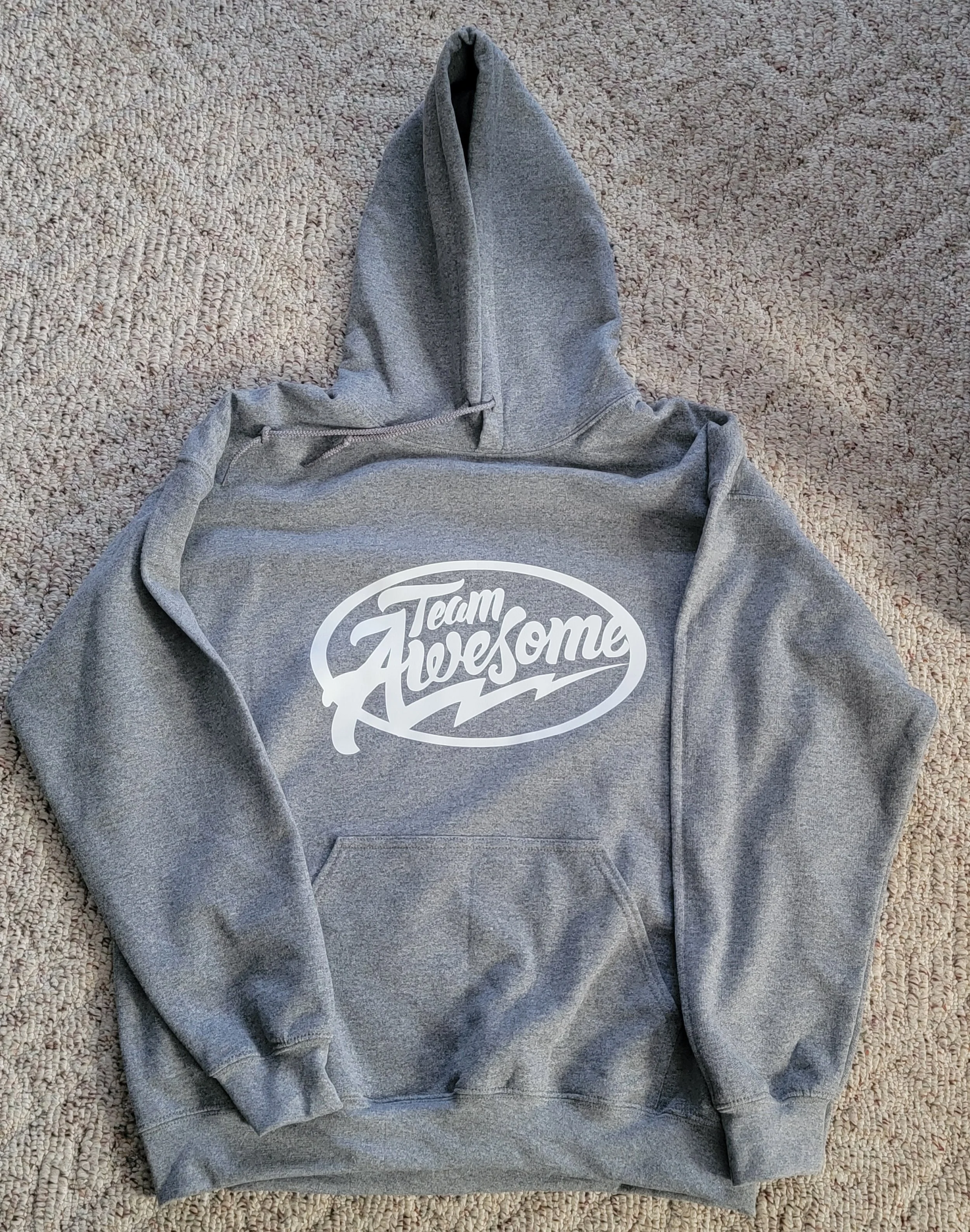 Gray hoodie with white 'Team Awesome' graphic on the front, laid flat on a beige carpet.