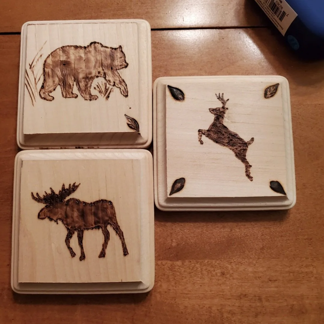 Three wooden boxes with images of a bear, a deer, and a moose burned or painted on the lids of each box, arranged on a wooden surface.