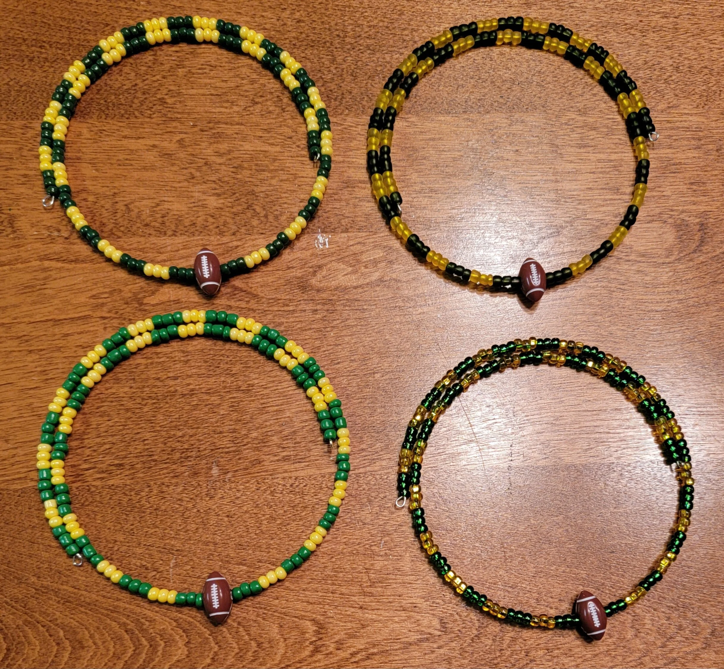 Four beaded necklaces with football-shaped pendants arranged on a wooden surface. Each necklace has a different color pattern: one with yellow and green beads, one with yellow and black beads, one predominantly green and yellow, and one with mixed bl