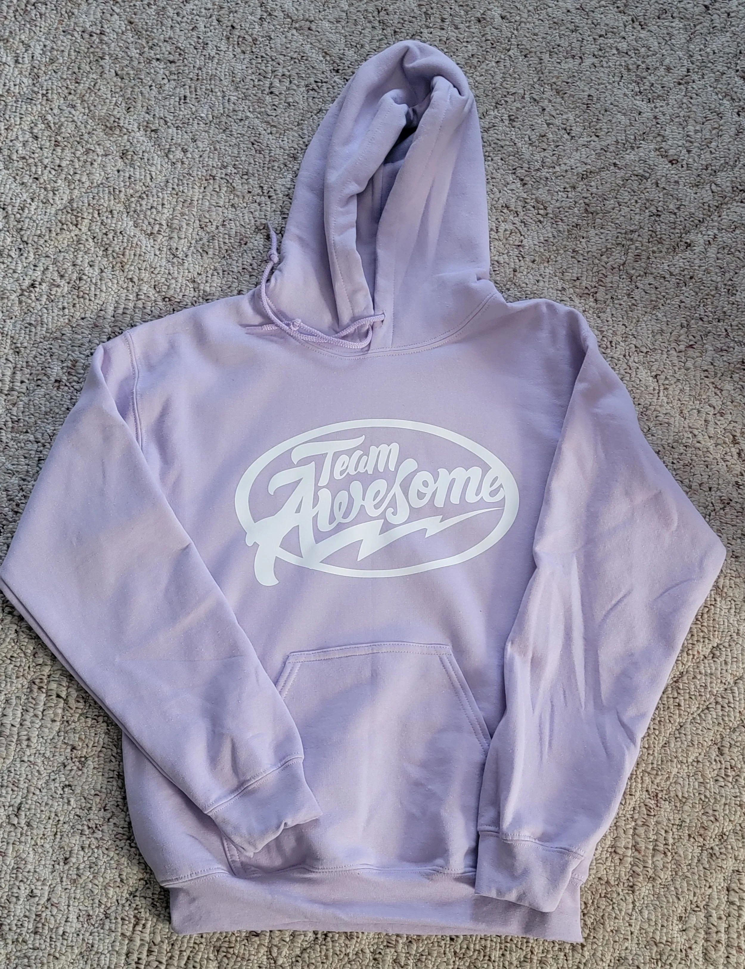 Light purple hoodie with a front pocket and drawstring hood, displaying 'Team Awesome' in white text inside an oval with lightning bolt accents, laid on a beige textured carpet.