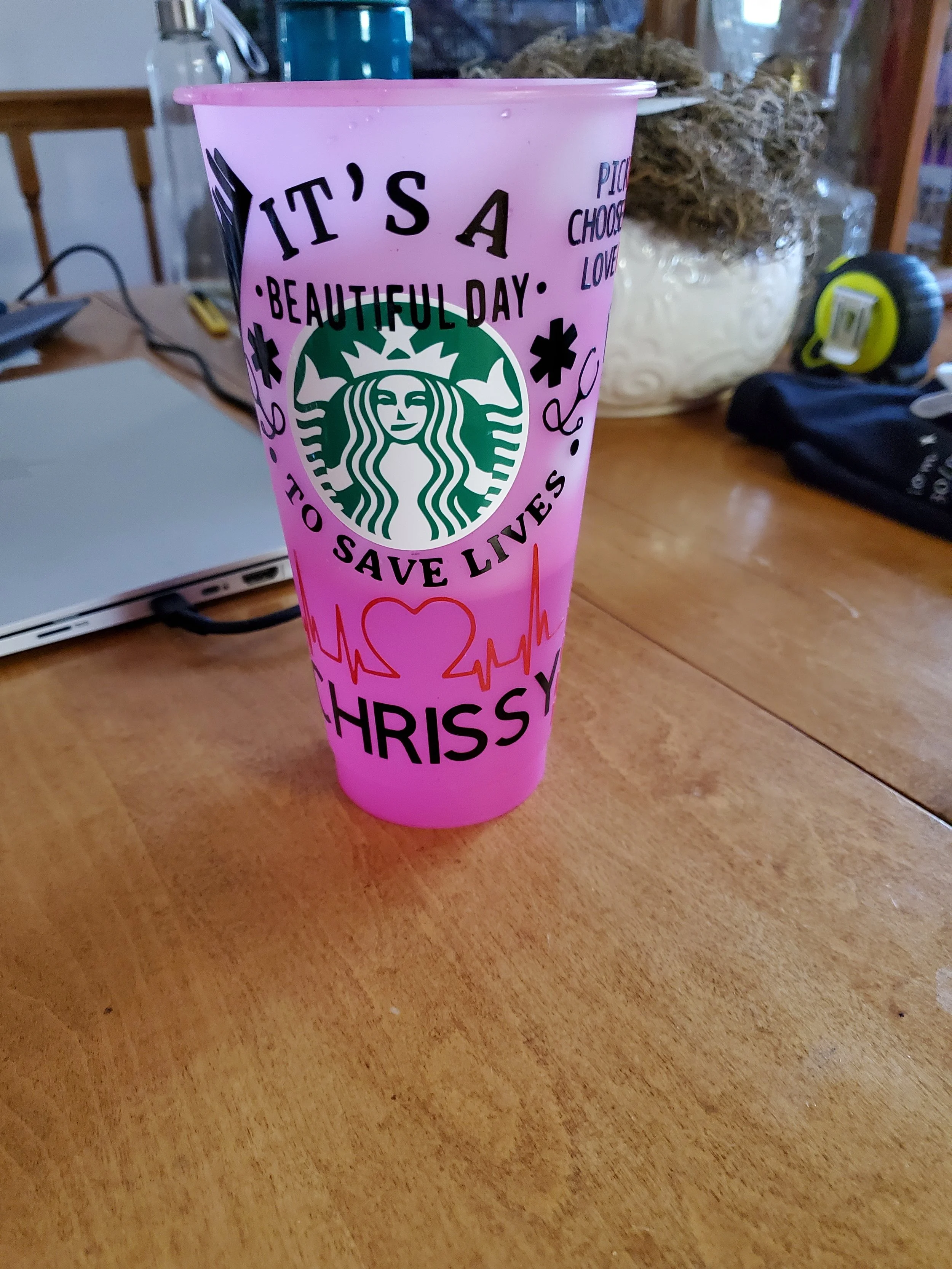 Pink Starbucks tumbler with the phrase 'It's a beautiful day to save lives' and 'Chrissy' written on it, placed on a wooden table with various items in the background.