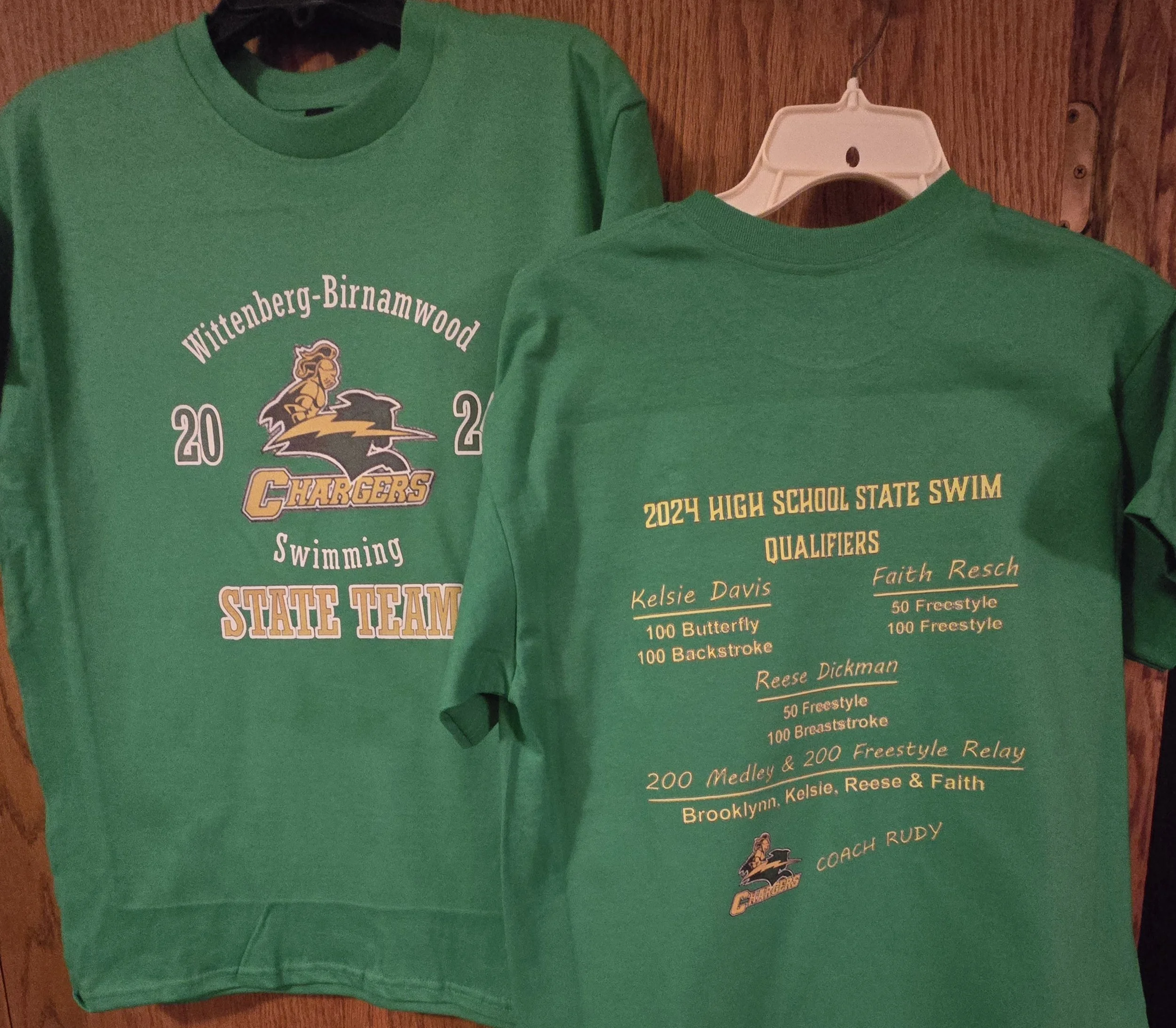Two green T-shirts hanging on a wooden wall, designed for a high school swim team. The front shirt has the school name 'Wittenberg-Birnamwood Chargers,' the year 2022, and 'Swimming State Team' printed on it. The back shirt displays information about