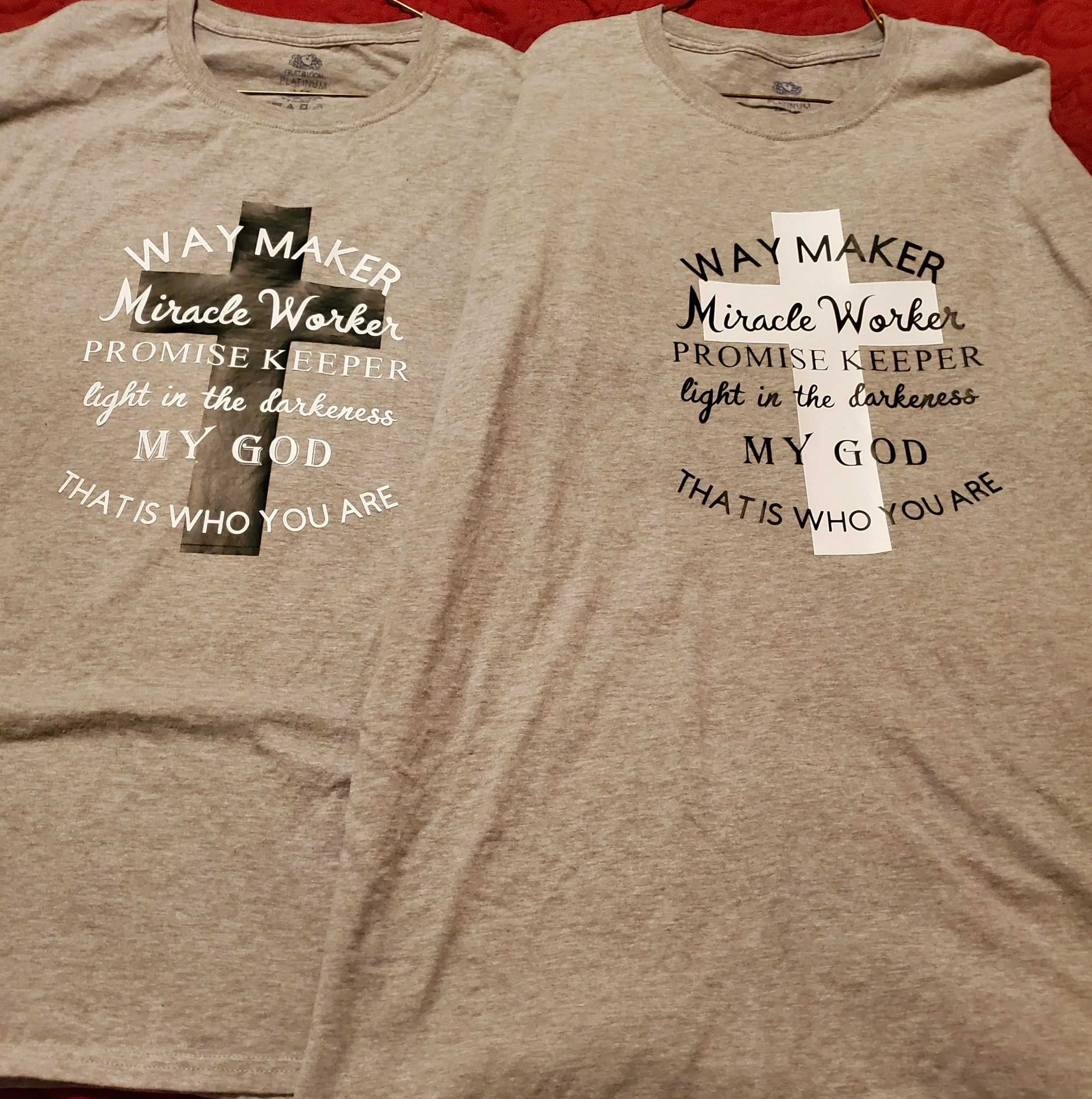 Two gray T-shirts side by side, each with a similar religious quote and a cross design. The left shirt has white text with a black cross, and the right shirt has black text with a white cross.