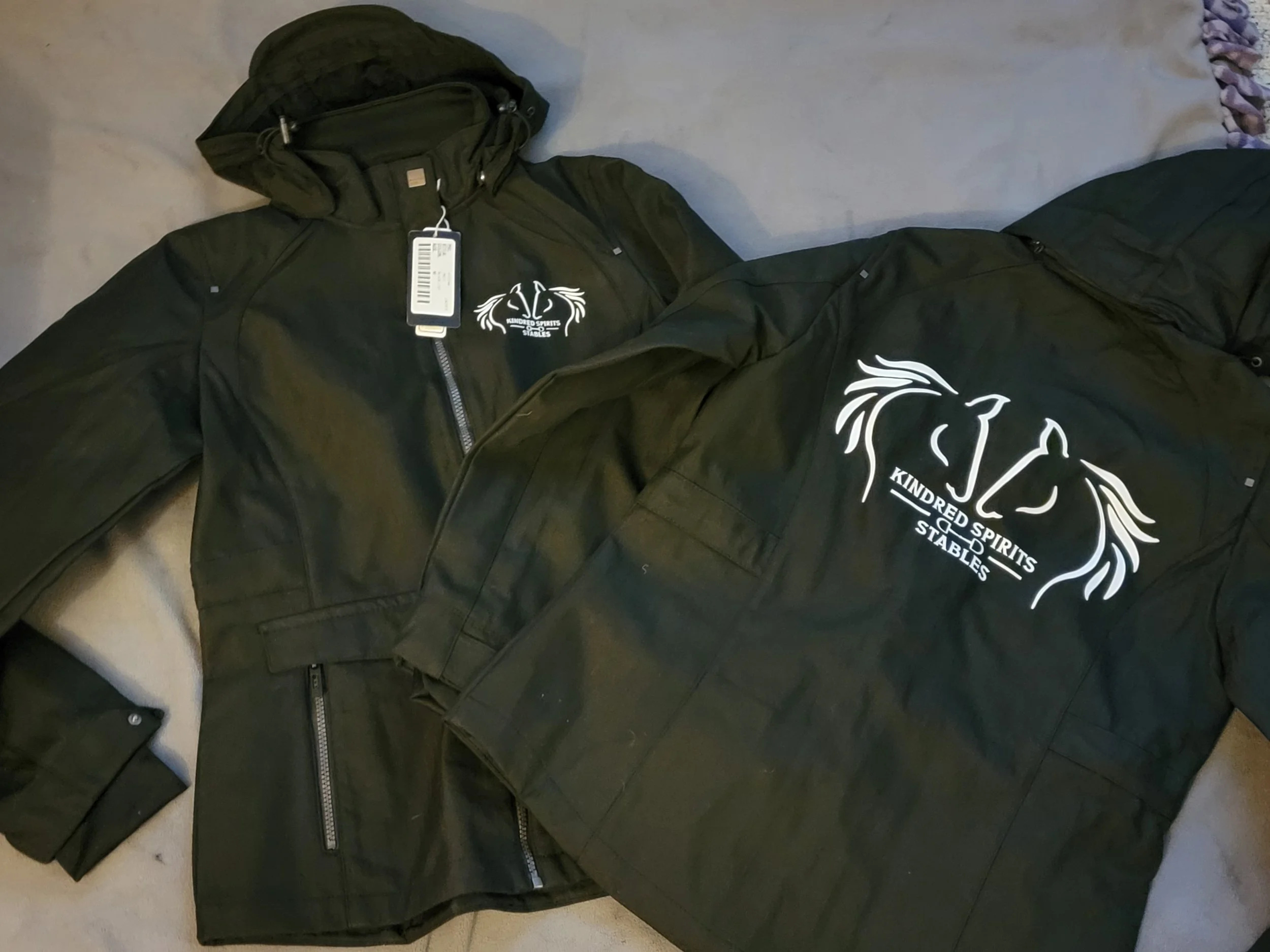 Two black jackets with a white logo and text that reads 'KINDRED SPIRITS STABLES' and small zippers, laid out on a grey surface.