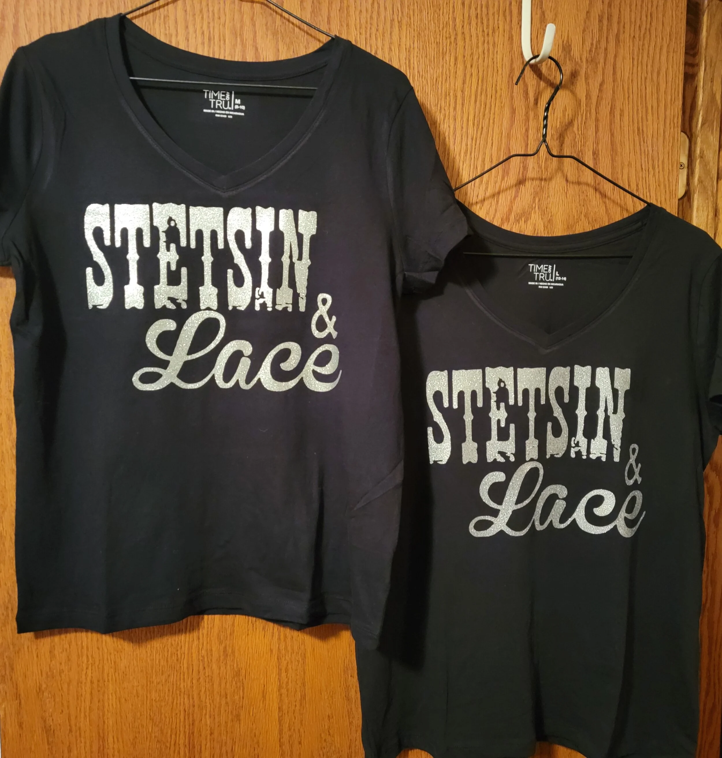 Two black T-shirts hanging on wooden wall background, with the text "Stetsin & Lace" in silver glitter font.
