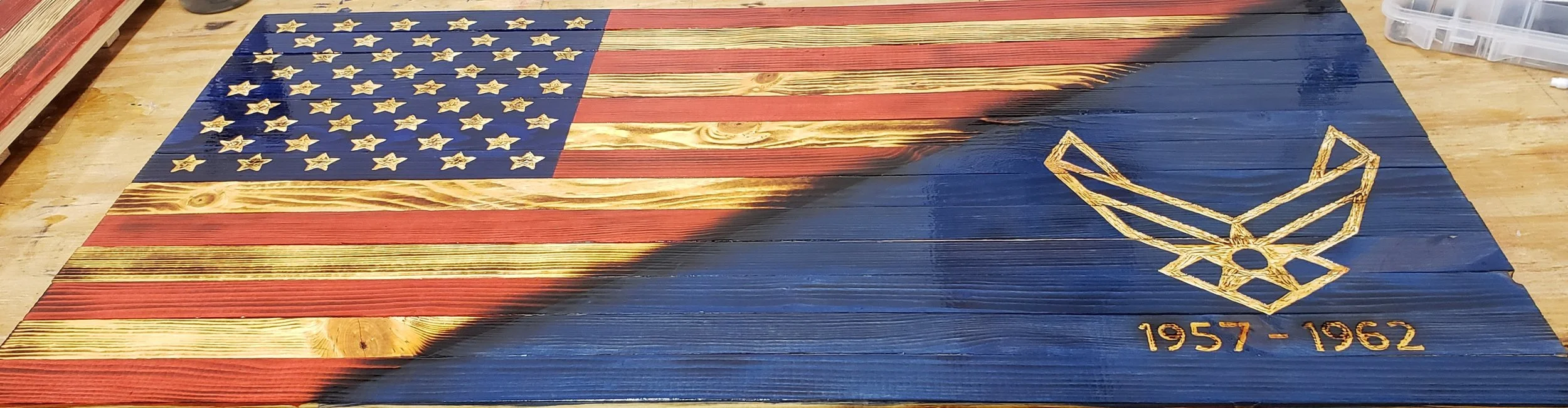 Wooden American flag with a blue canton with stars and red and wood stripes, partially painted with black. Golden emblem and years "1957-1962" are on the bottom right.