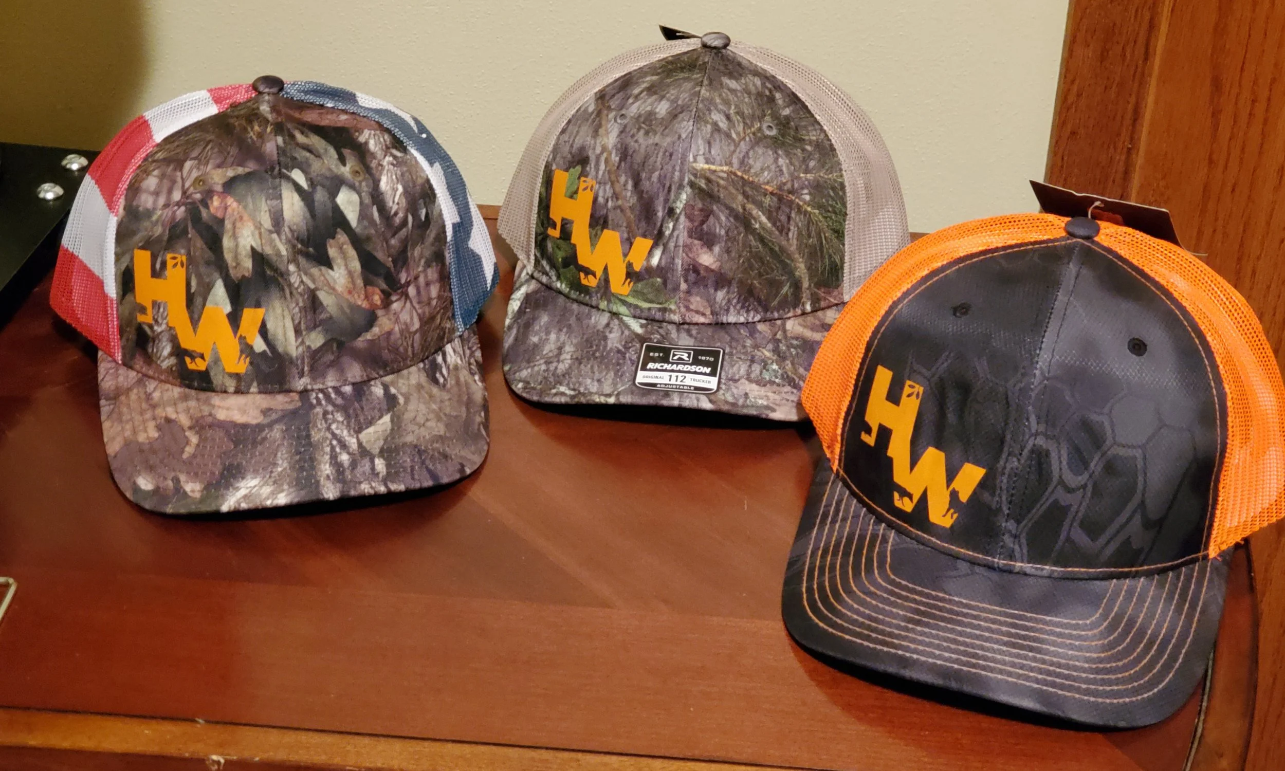 Three trucker hats with camouflage and black logos placed on a wooden surface. The hats feature different mesh colors—red, beige, and orange—and the same logo on the front.