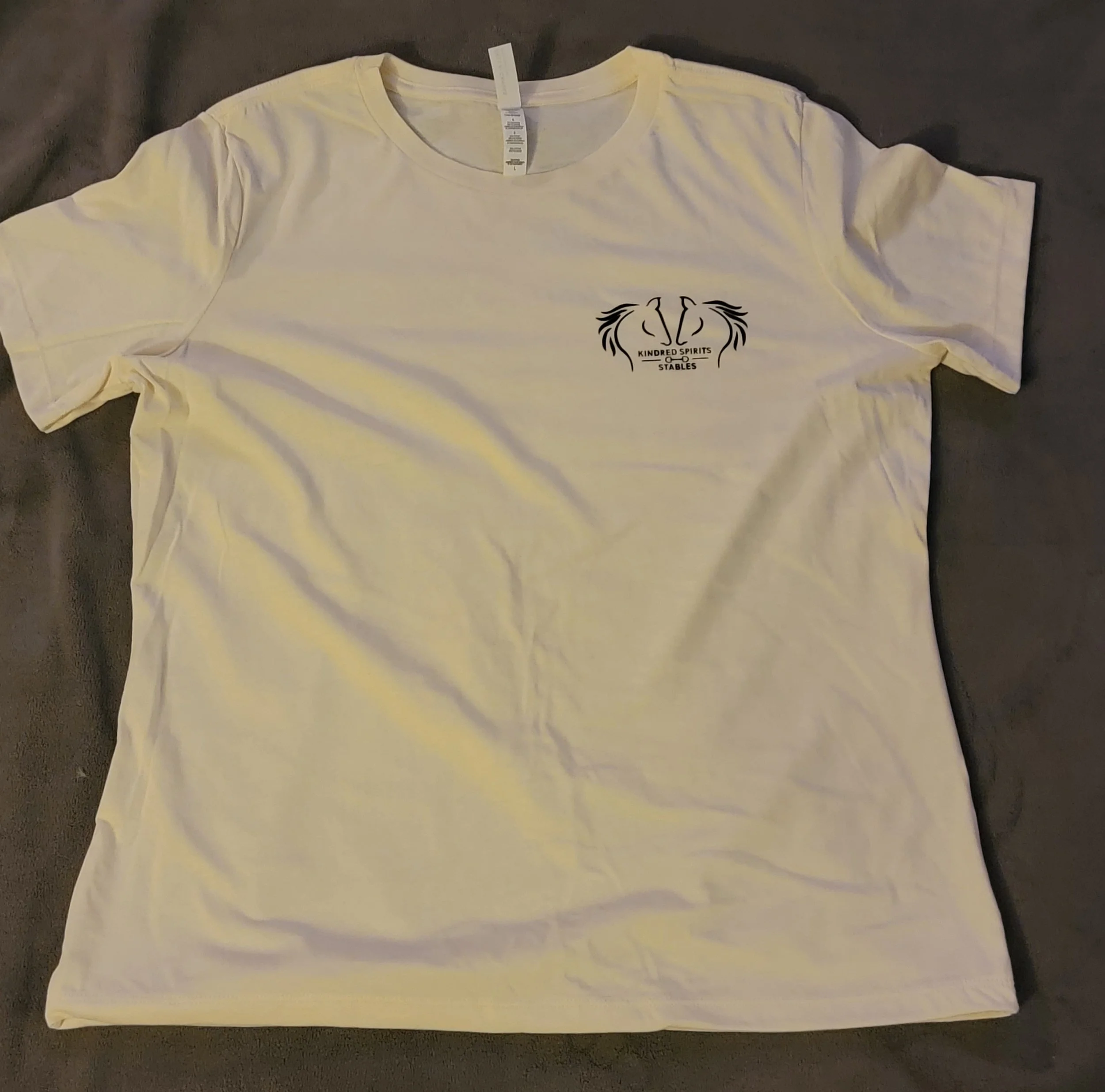 A white T-shirt with a small black logo on the left chest area. The logo features a stylized horse head outline with the text 'KINDRED SPIRITS STABLES' inside it.