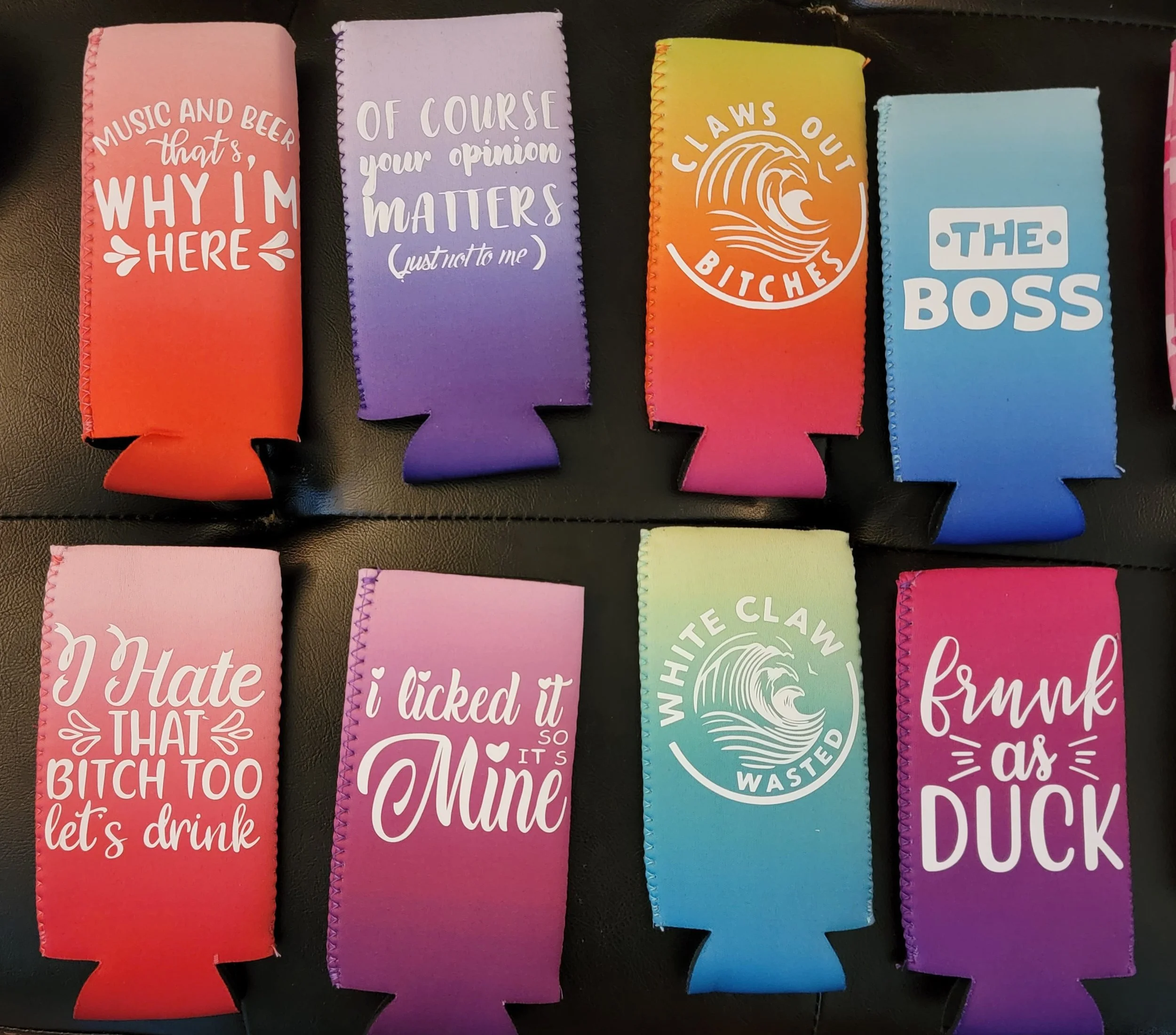Colorful foam drink koozies with humorous and sassy phrases written in white text, arranged in two rows on a black surface.