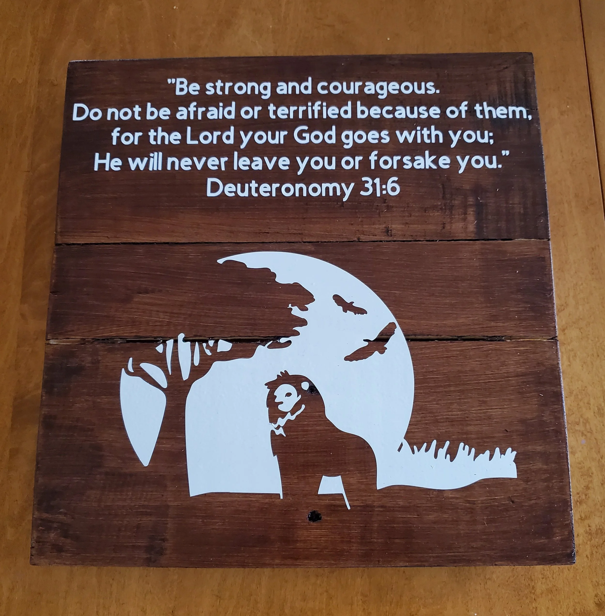 A wooden plaque with a quote from Deuteronomy 31:6 and a cutout silhouette of a lion, tree, grass, and flying birds against a moon or sun in the background.