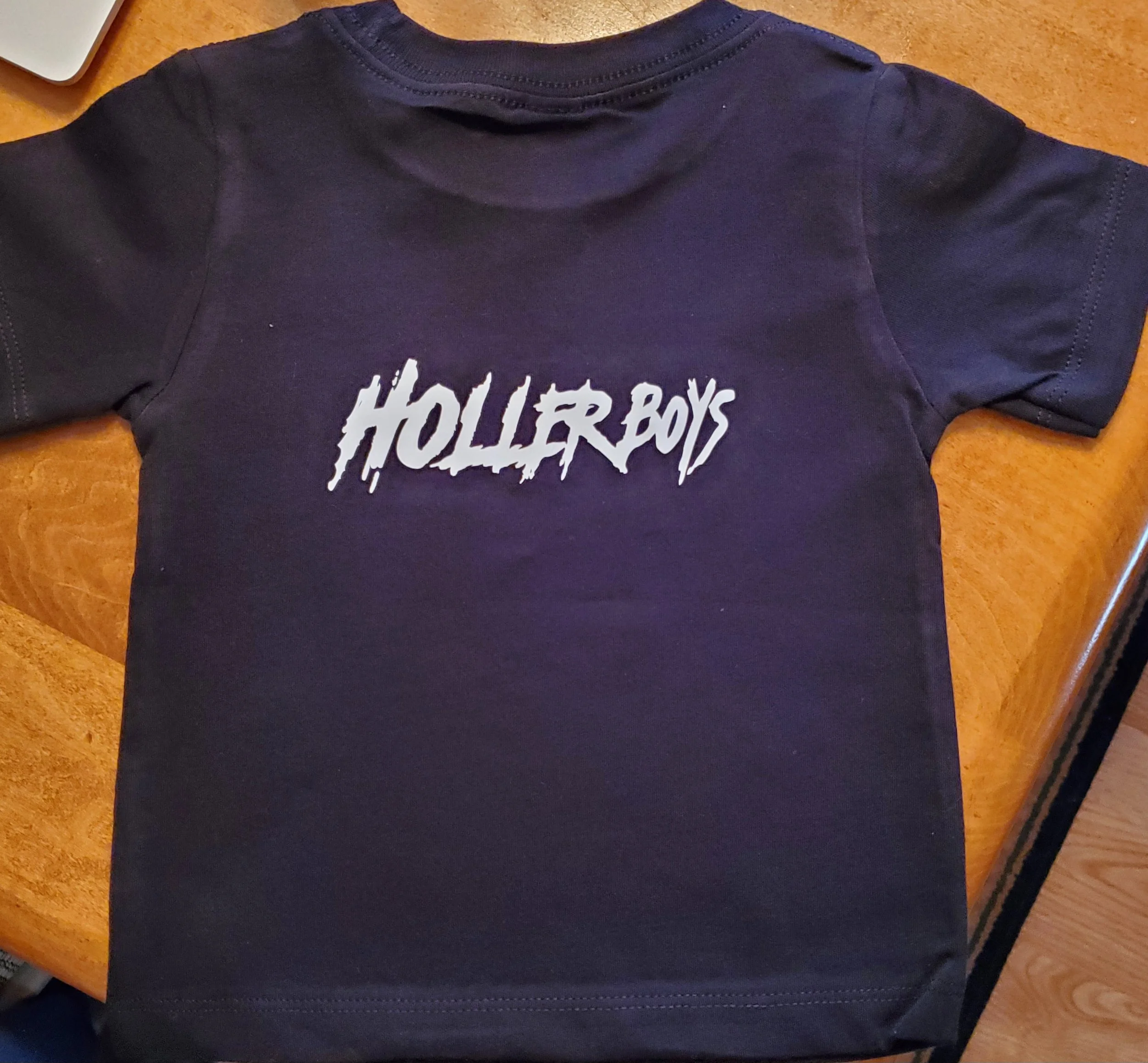 Black short-sleeve t-shirt with white 'Holler Boys' text logo on the front, lying on a wooden surface.