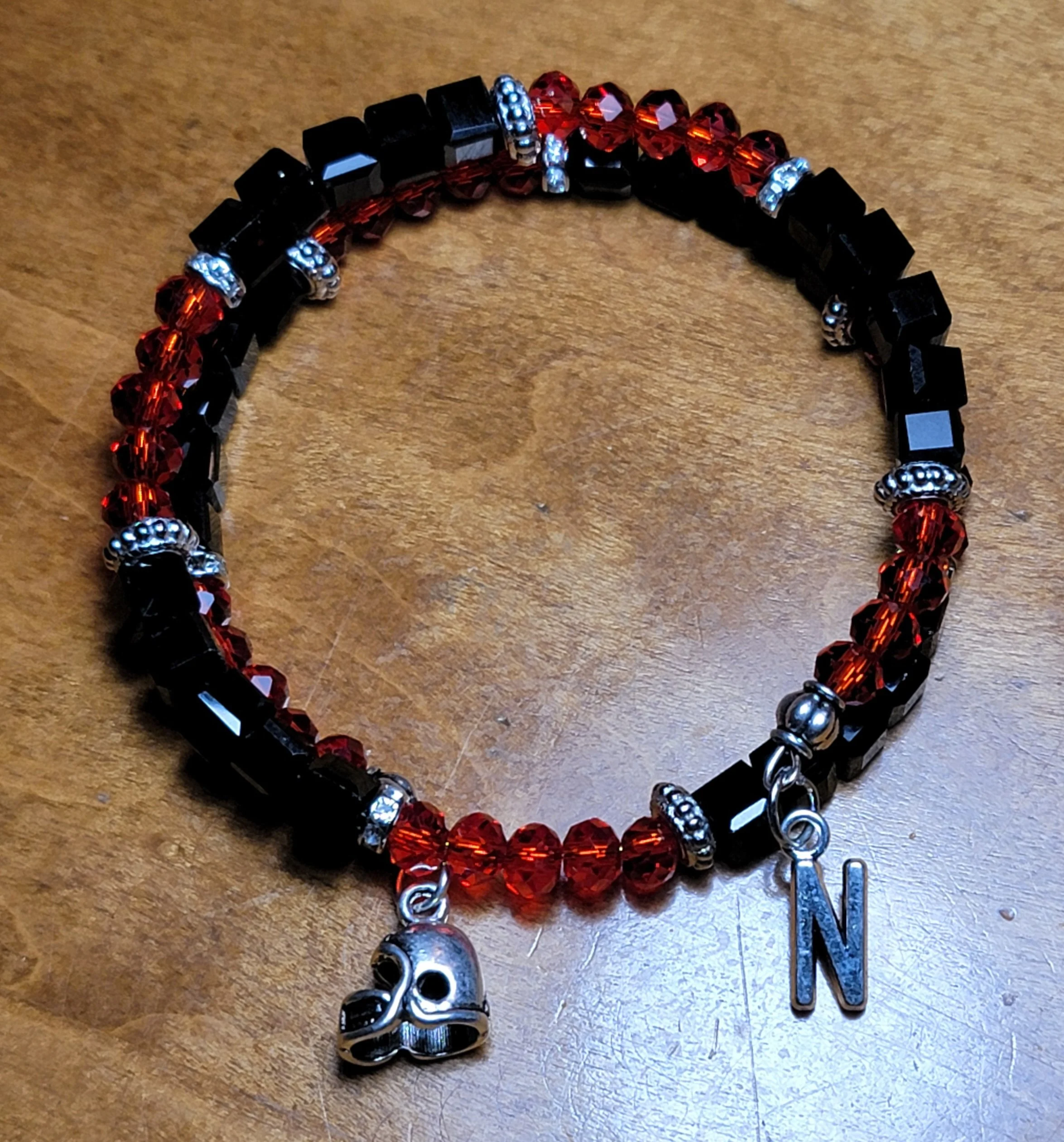 Beaded bracelet with red and black beads, silver skull charm, and a silver 'N' charm, placed on a wooden surface.