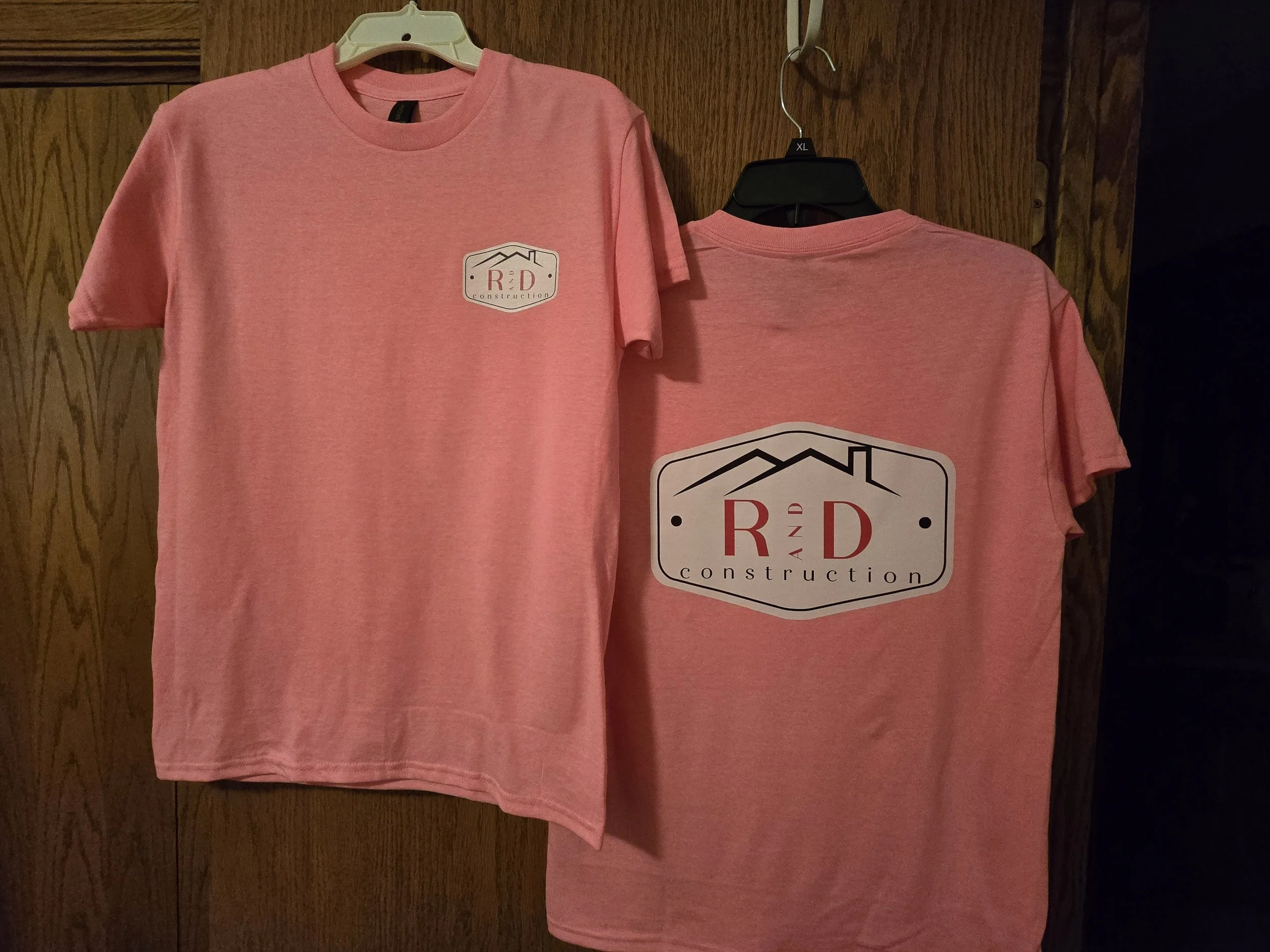 Pink T-shirts with a logo for R&D Construction hanging on a wooden wall.