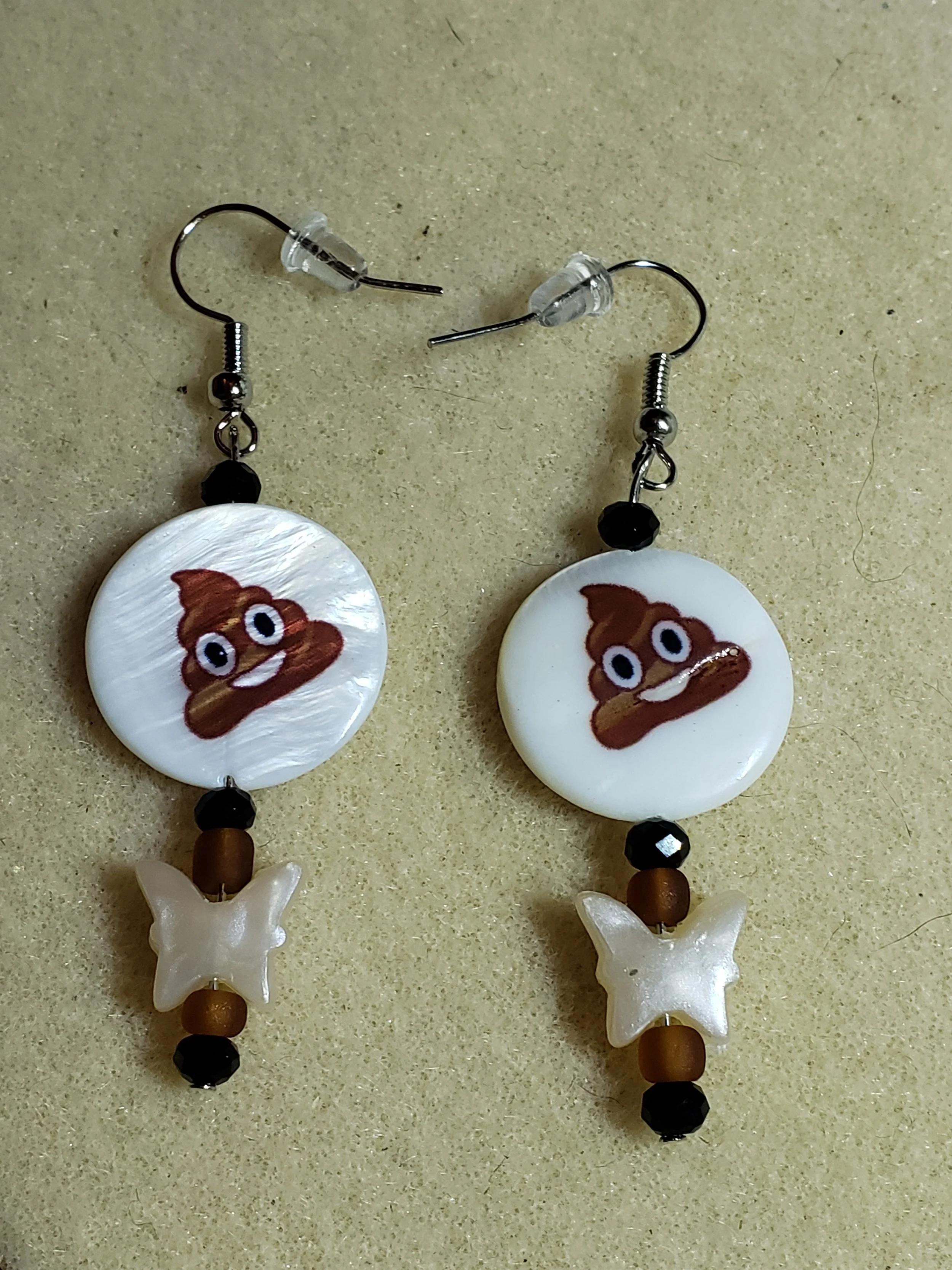 Pair of earrings with a poop emoji graphic, black and brown beads, and white butterfly charms, on a beige surface.