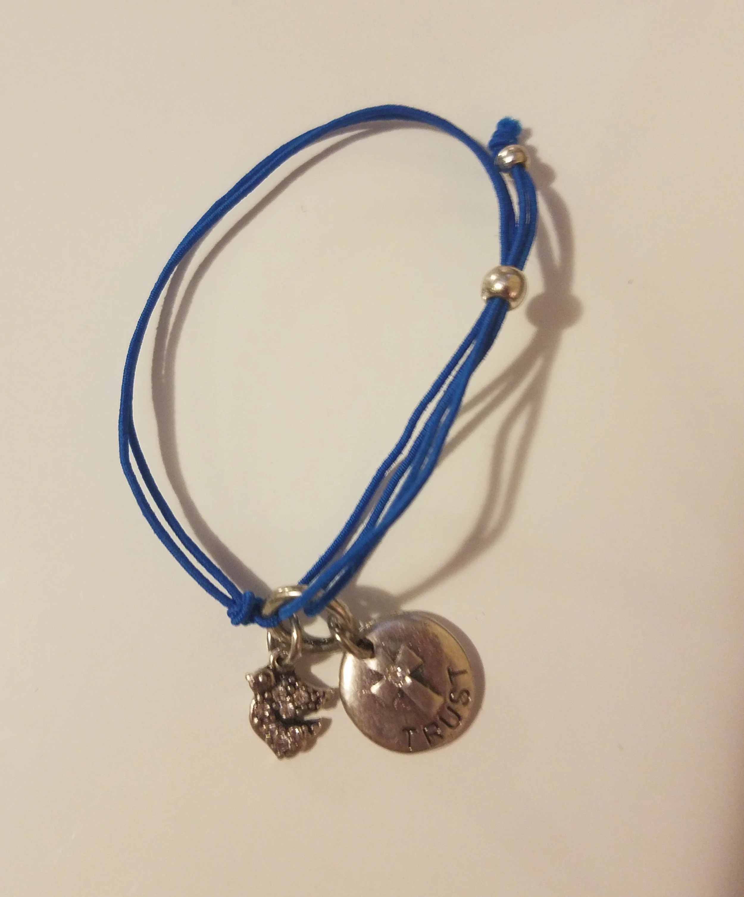 Blue string bracelet with three charms: a peace sign, a round disc with a cross and the word 'TRUST', and a small angel or fairy figure with rhinestones.