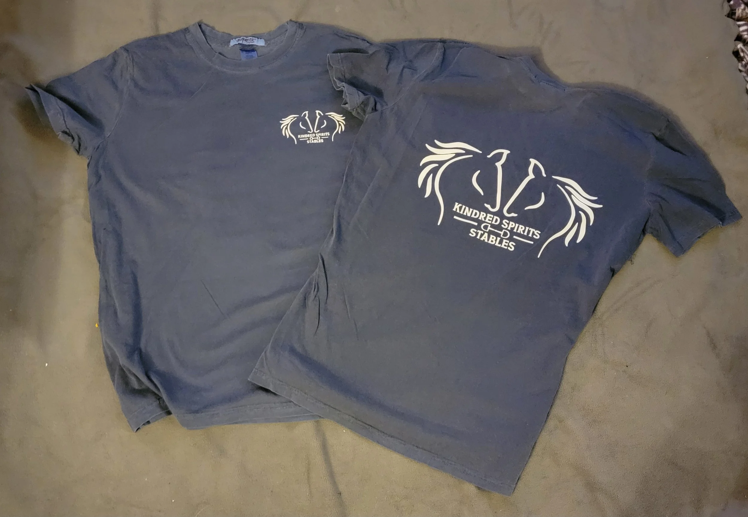 Two gray T-shirts laid flat on a surface, both featuring a white graphic design on the front and back that depicts a stylized horse's head and the text 'KINDRED SPIRITS STABLES'.