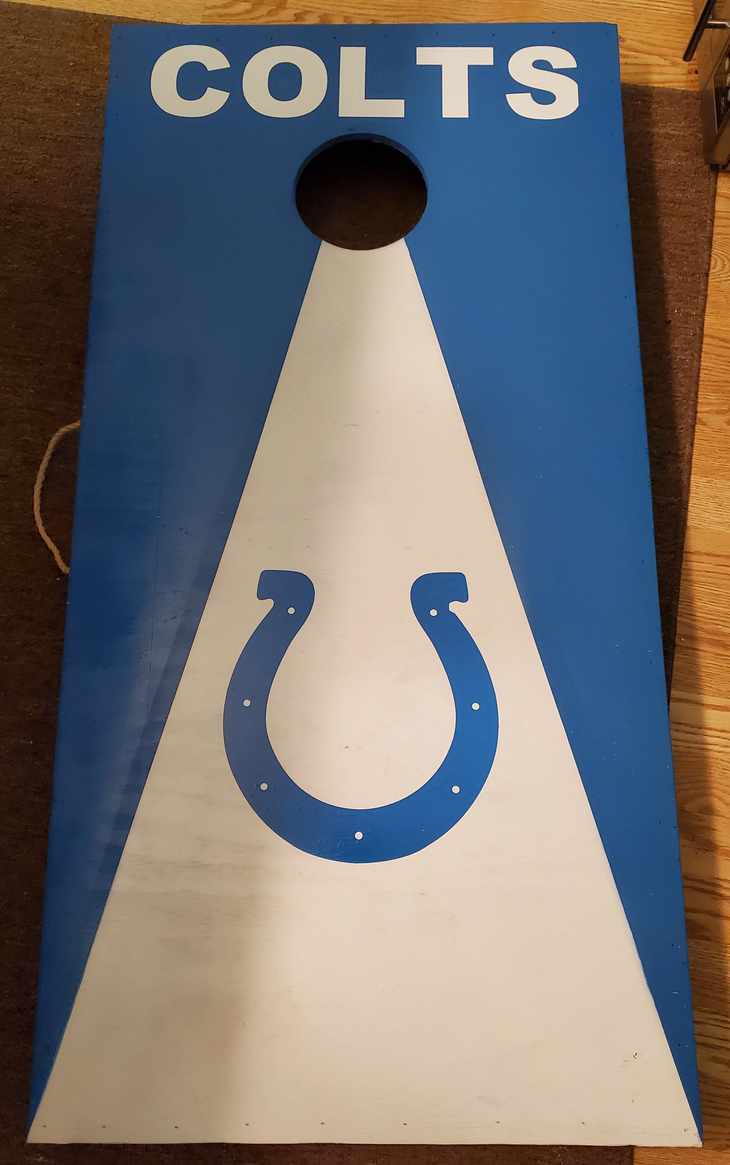 A horseshoe game toss board painted in blue and white with the word 'COLTS' at the top. It has a circular hole near the top center and a horseshoe in the bottom center, with a white lightning bolt-shaped design connecting the hole and the horseshoe.