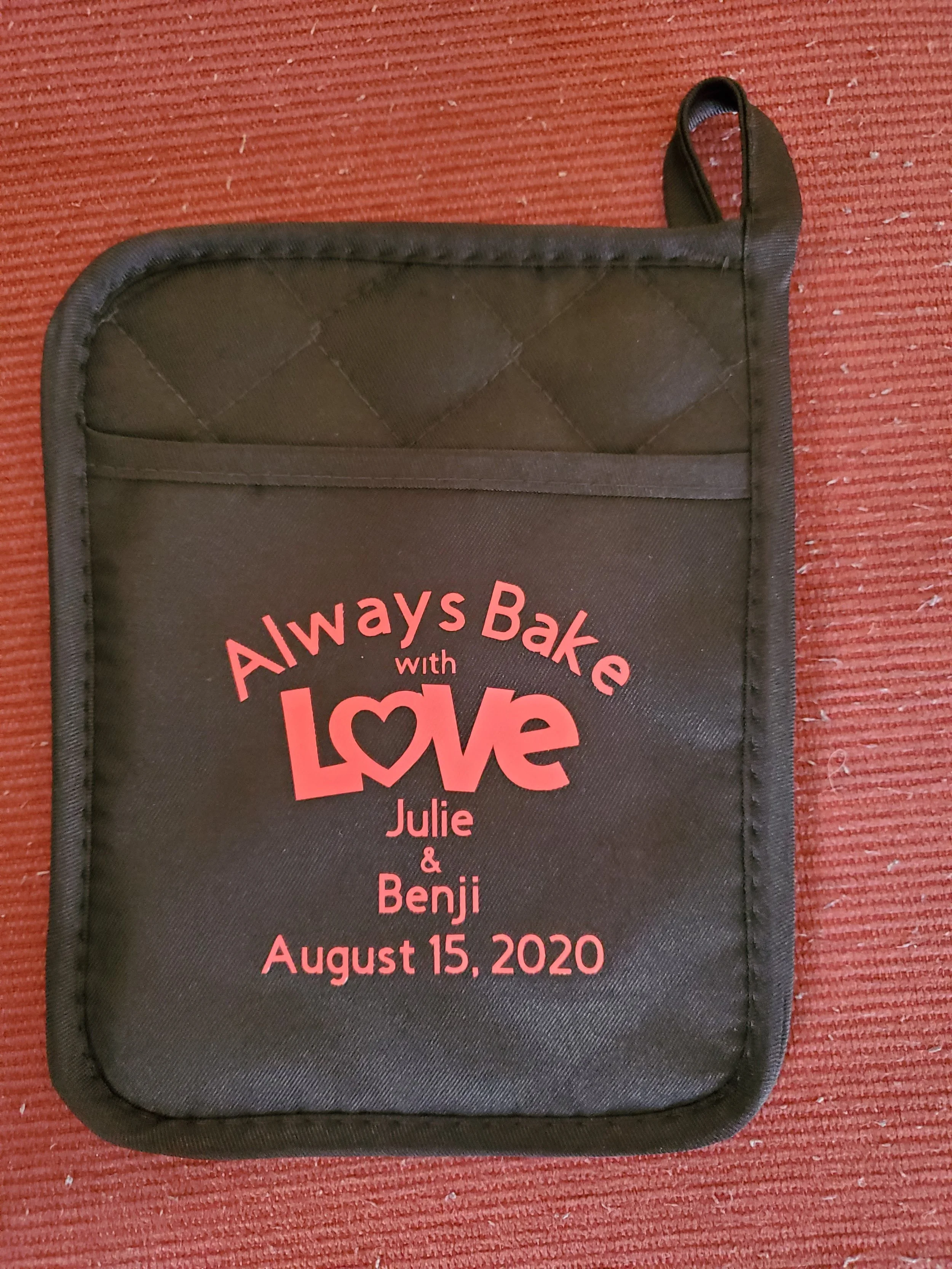 Black oven mitt with red text that reads 'Always Bake with Love, Julie & Benji, August 15, 2020' on a red textured surface.