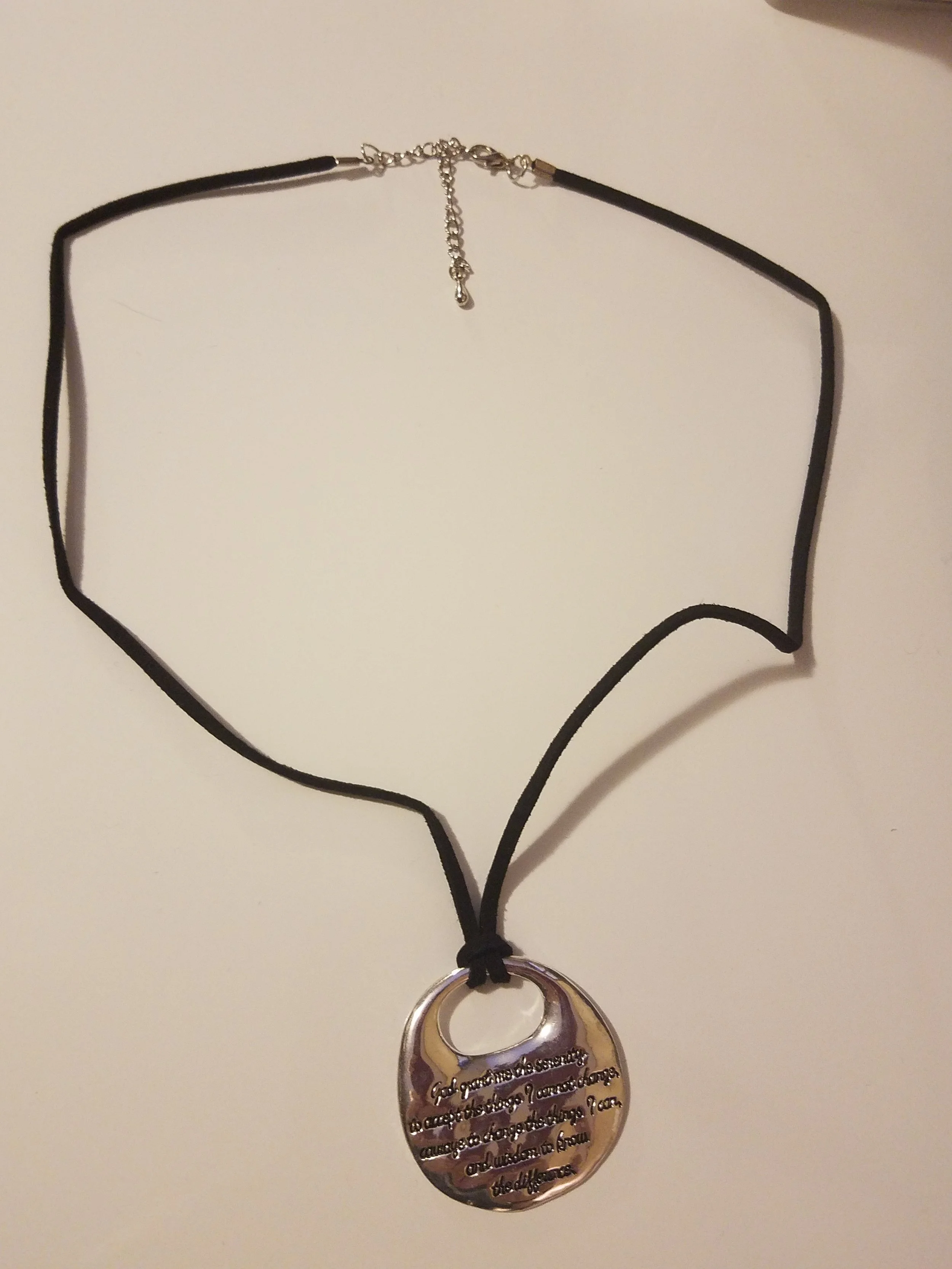 A silver pendant necklace with an inscription hanging from a black cord, laid on a white surface.