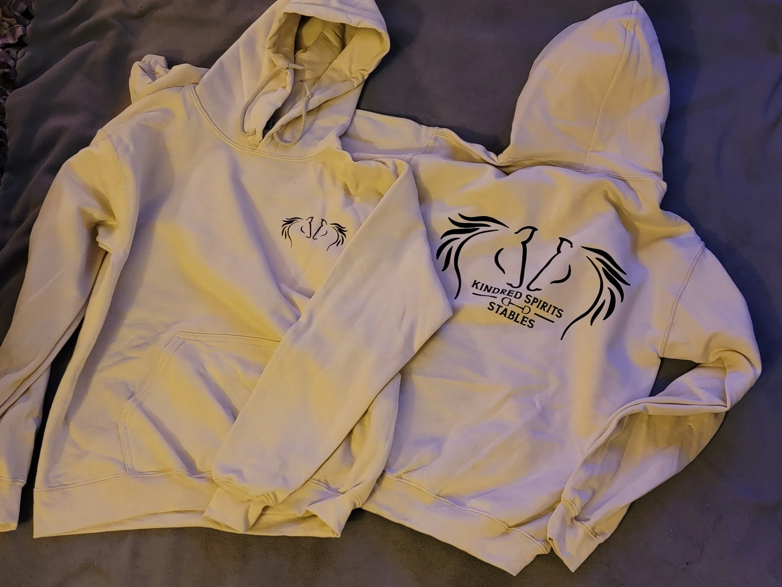 Two cream-colored hoodies with black horse and mane logo and text that reads 'KINDRED SPIRITS STABLES' on the back are laid flat on a dark surface.