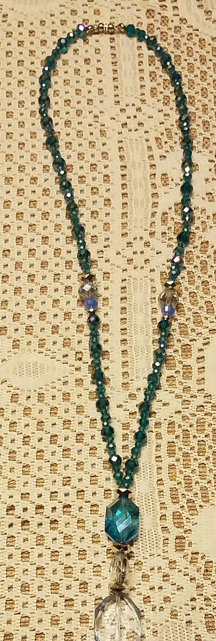 Long beaded necklace with a large blue gemstone pendant, resting on a beige textured fabric background.