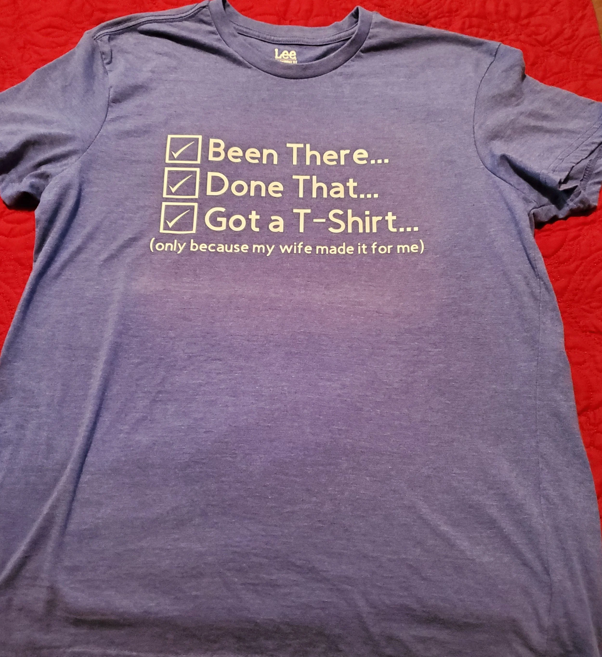 A purple T-shirt with text that reads, 'Been There... Done That... Got a T-Shirt...' with checkboxes next to each phrase, and a note underneath stating, '(only because my wife made it for me)'.