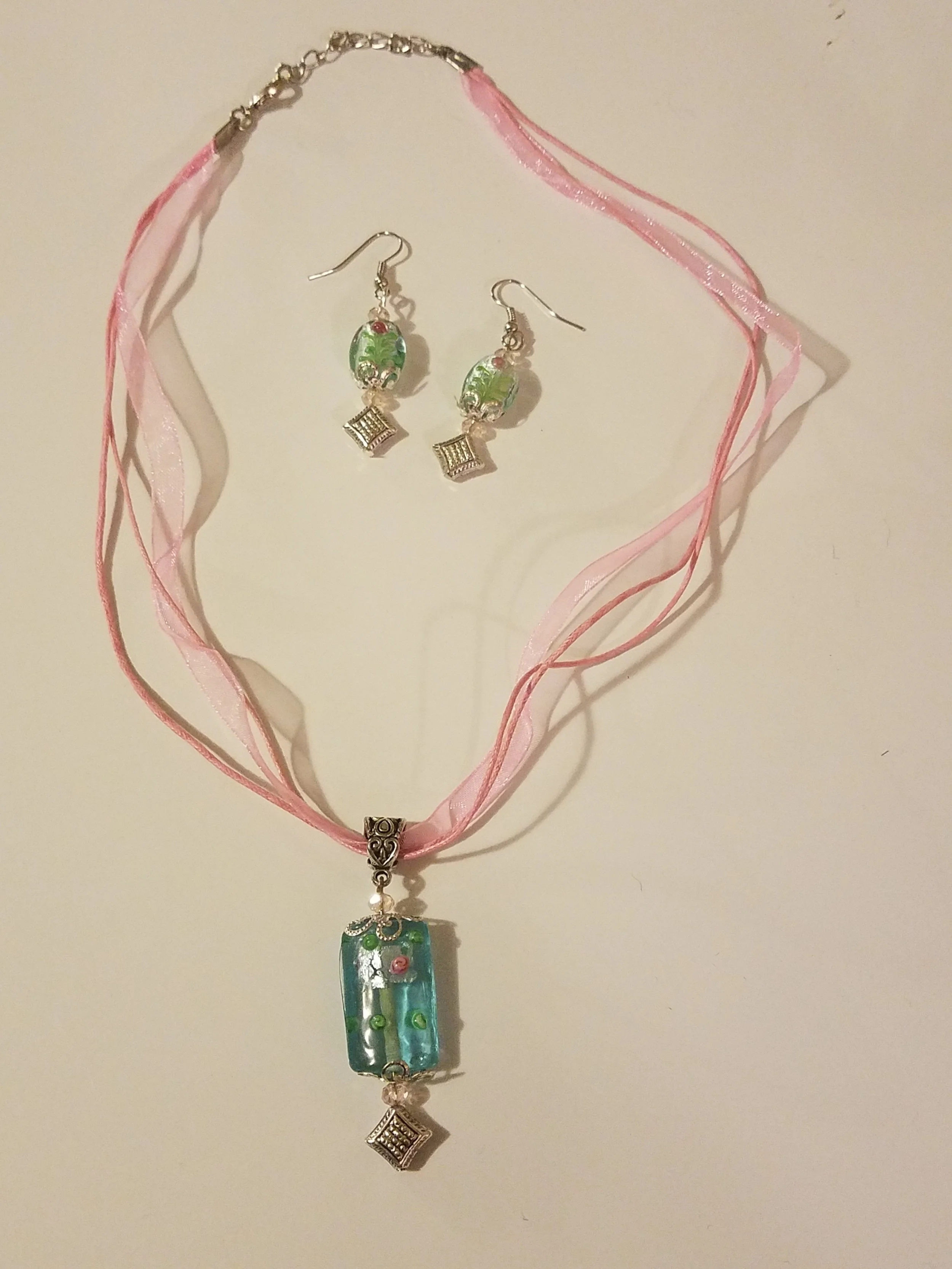 Necklace and earrings set featuring green and clear beads with silver accents. The necklace has a pink ribbon-like chain with a rectangular pendant. The earrings have oval beads and a square silver charm.