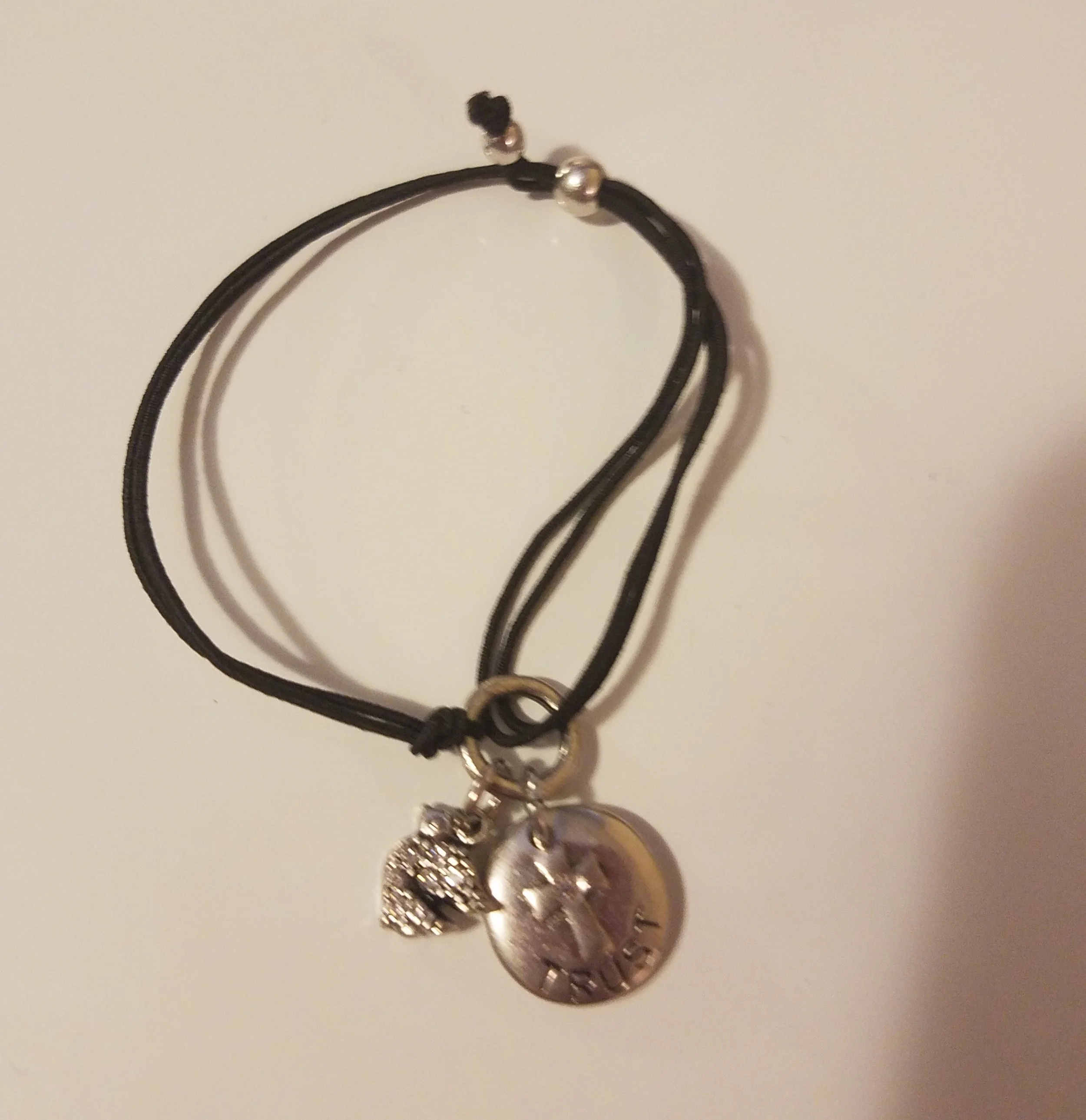 A black cord bracelet with three charms, including a small owl, a round silver coin, and a heart.